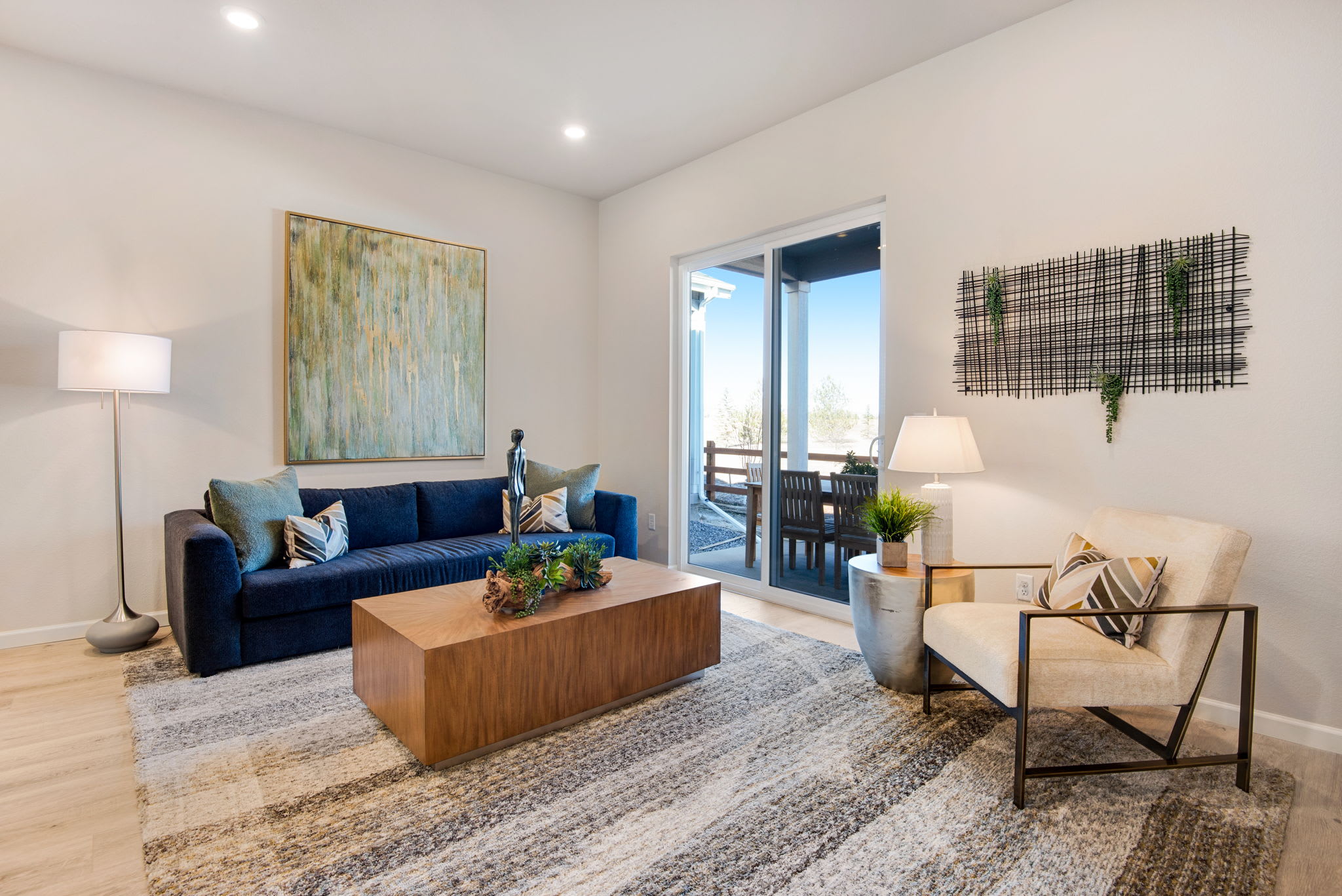 A cozy and inviting living room with a navy blue sofa, a wooden coffee table, and various decorative elements such as artwork, plants, and lamps, all set against a bright and airy background with a view of the outdoors.
