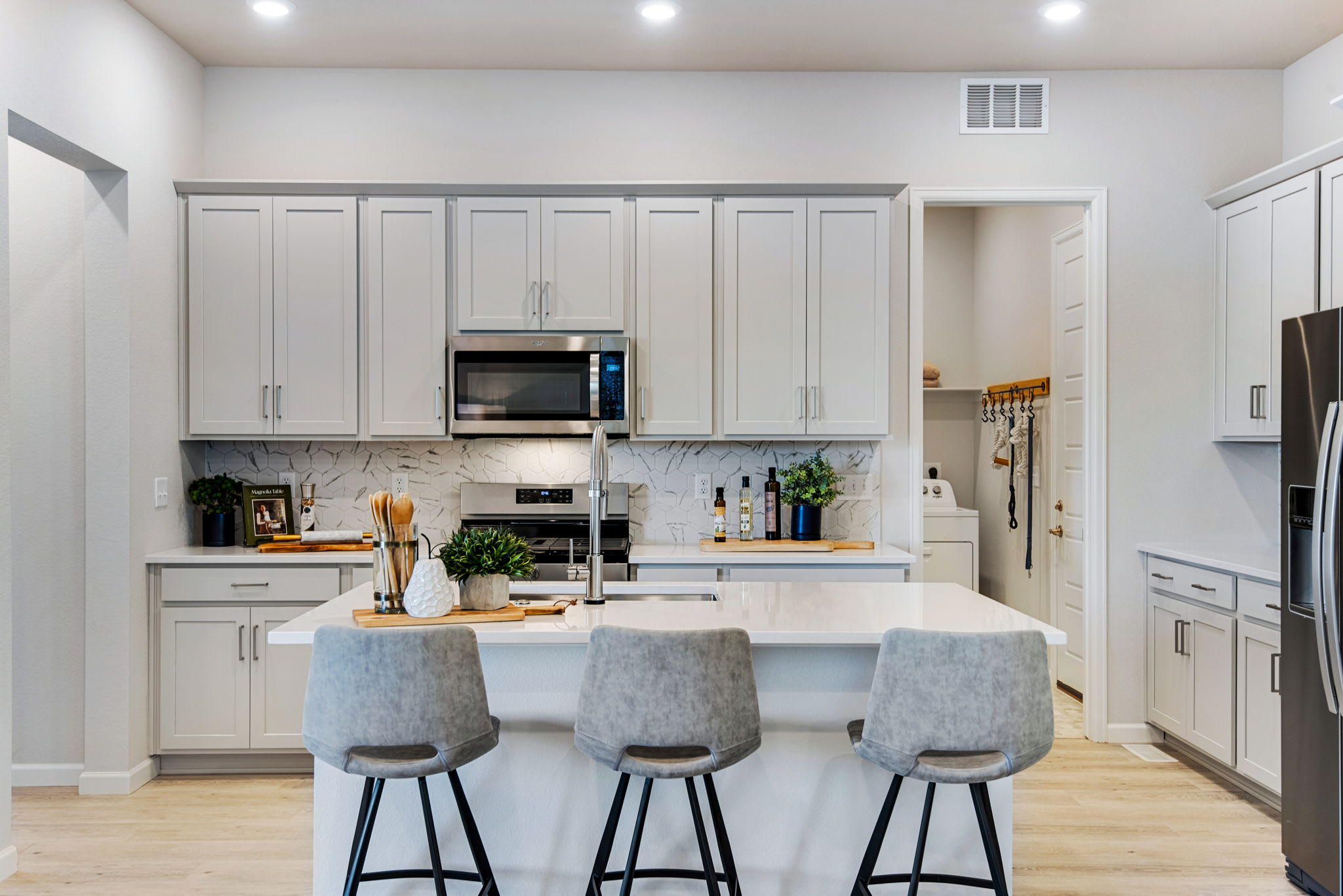 A modern, well-equipped kitchen with white cabinets, a central island, and gray bar stools, set against a light-colored hardwood floor.