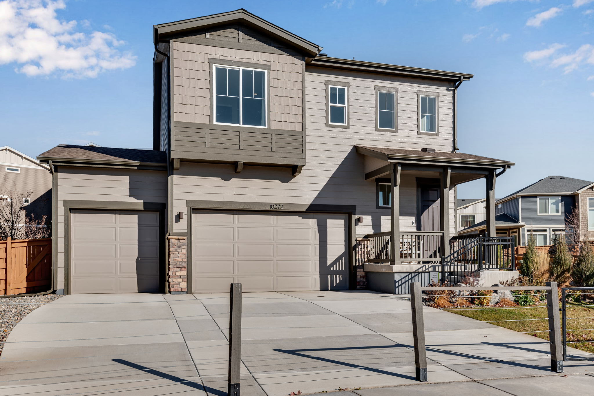 A modern two-story residential house with a garage, surrounded by a paved driveway and a well-maintained yard with landscaping in the foreground, set against a clear blue sky with scattered clouds.