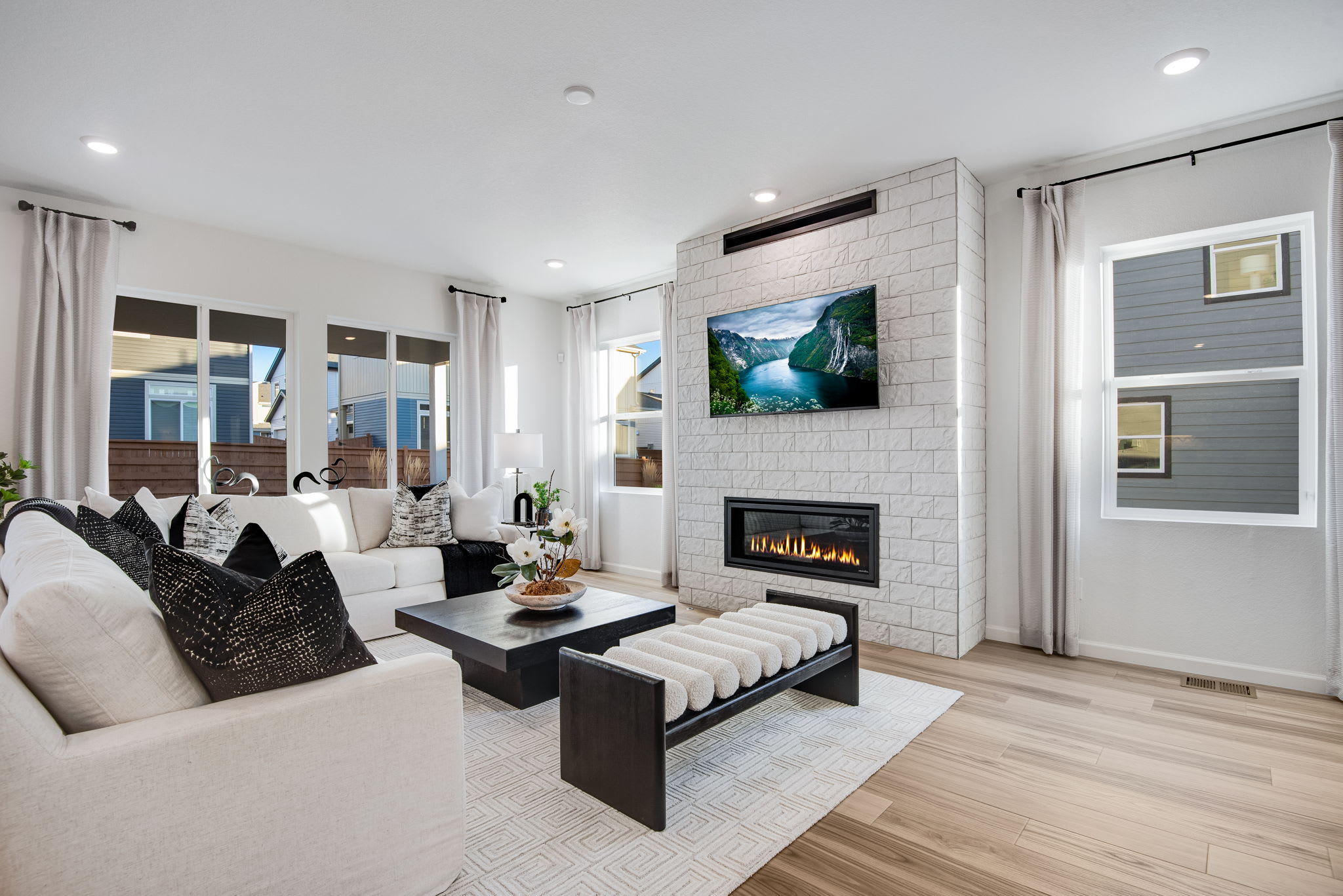 A modern and cozy living room with a fireplace, large windows, and a stylish furniture arrangement featuring a white sofa, black coffee table, and a vibrant landscape artwork on the wall.
