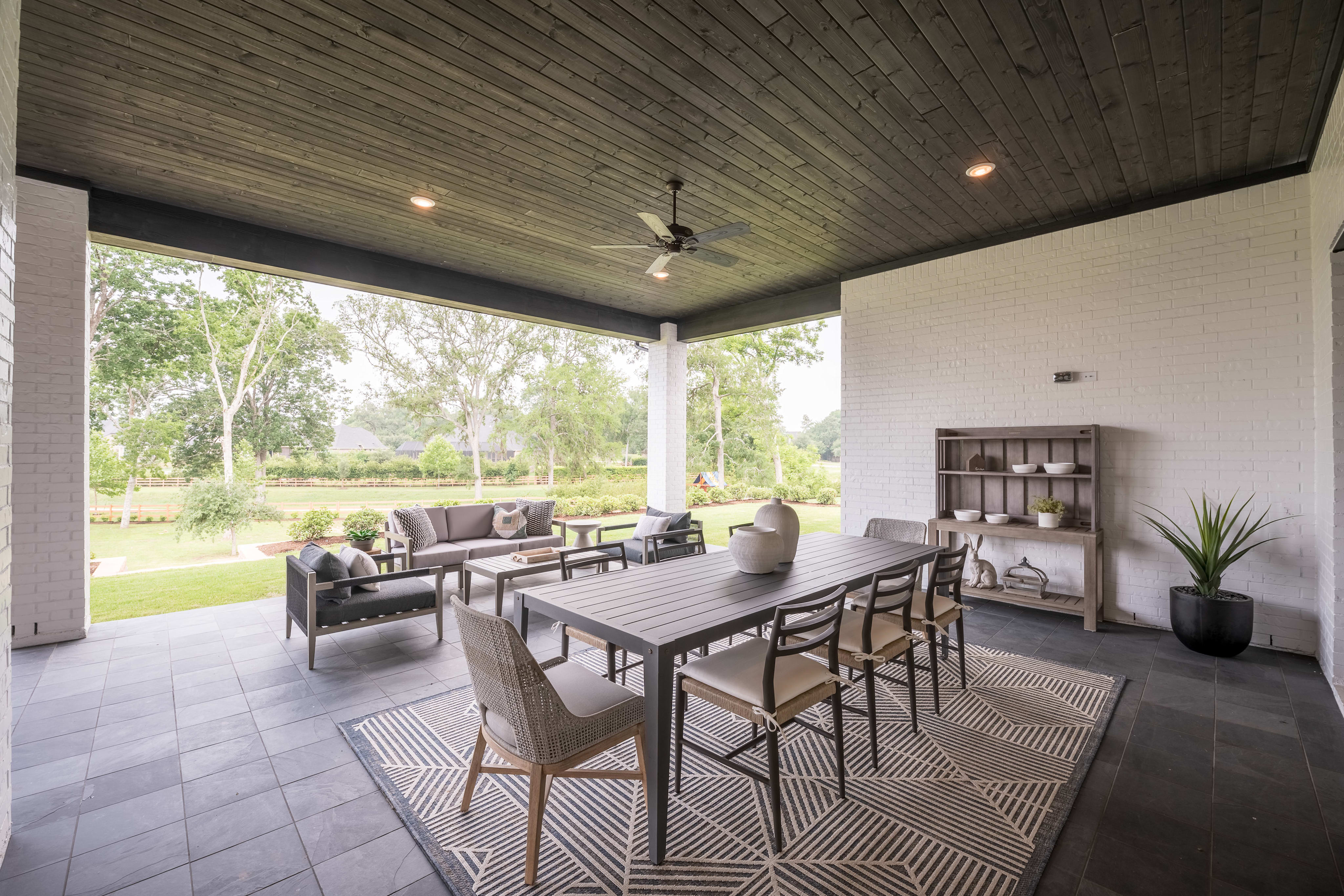 A covered outdoor patio with a dining table and chairs, surrounded by lush greenery and trees visible through the large windows.