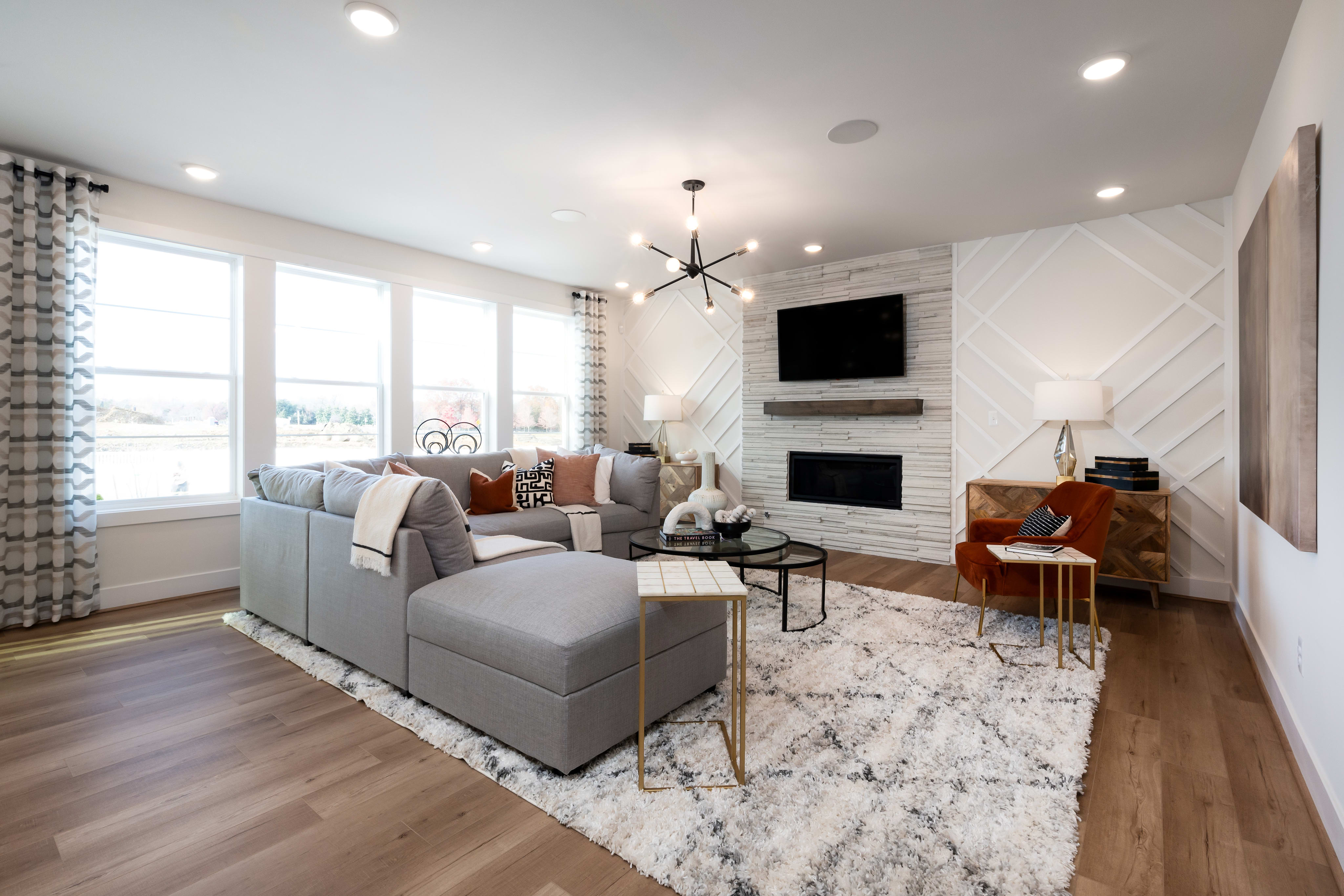 A cozy and modern living room with a gray sectional sofa, a patterned area rug, and a fireplace with a mounted TV above it, surrounded by large windows that let in natural light.