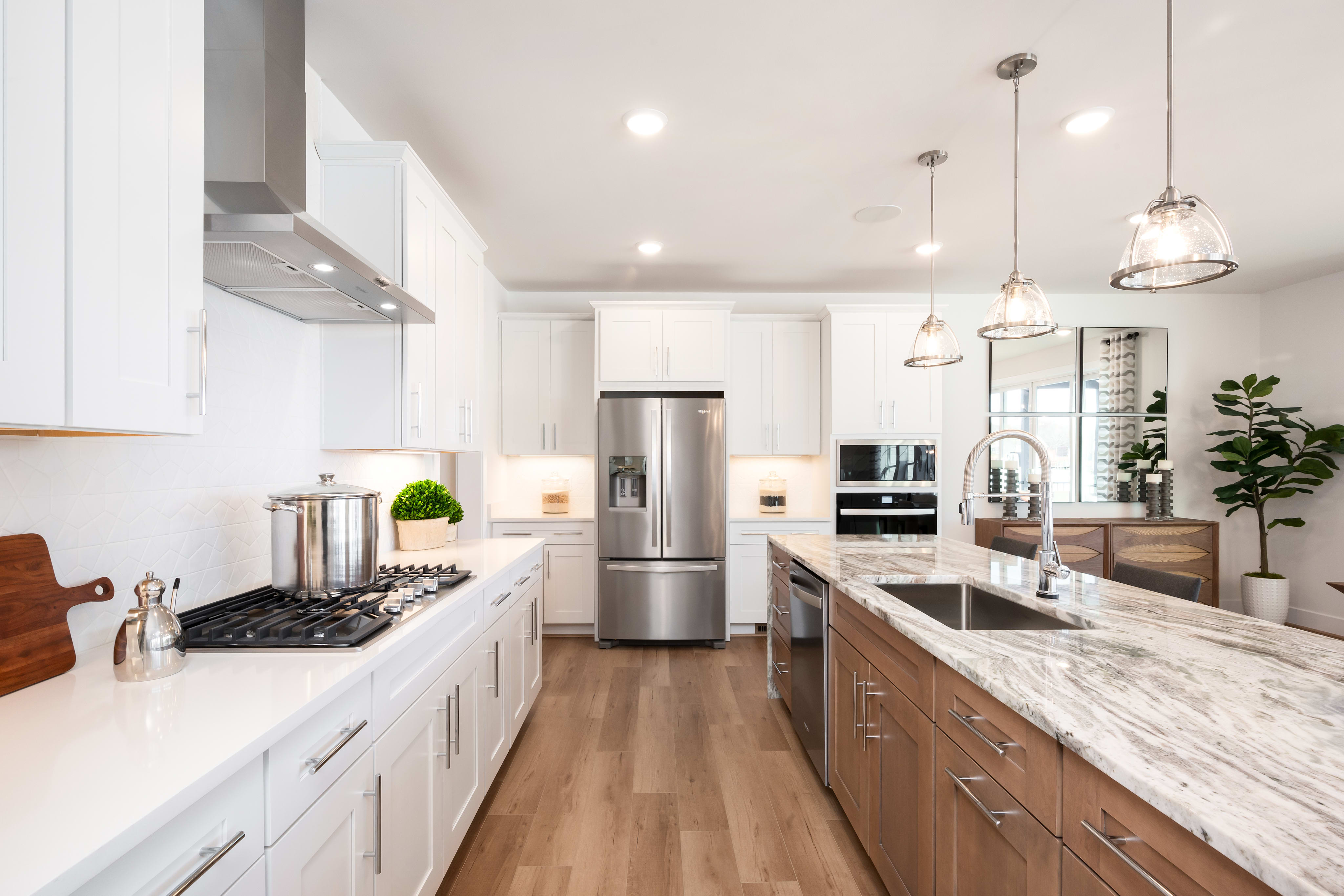 A modern, well-lit kitchen with white cabinets, marble countertops, and stainless steel appliances, featuring a long central island and pendant lighting.