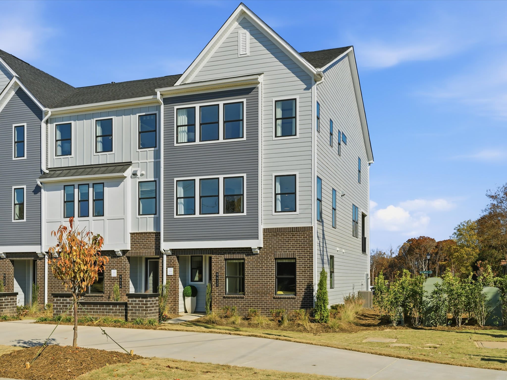 A multi-story townhouse with a brick and siding exterior, surrounded by landscaping and a paved walkway, set against a clear blue sky.