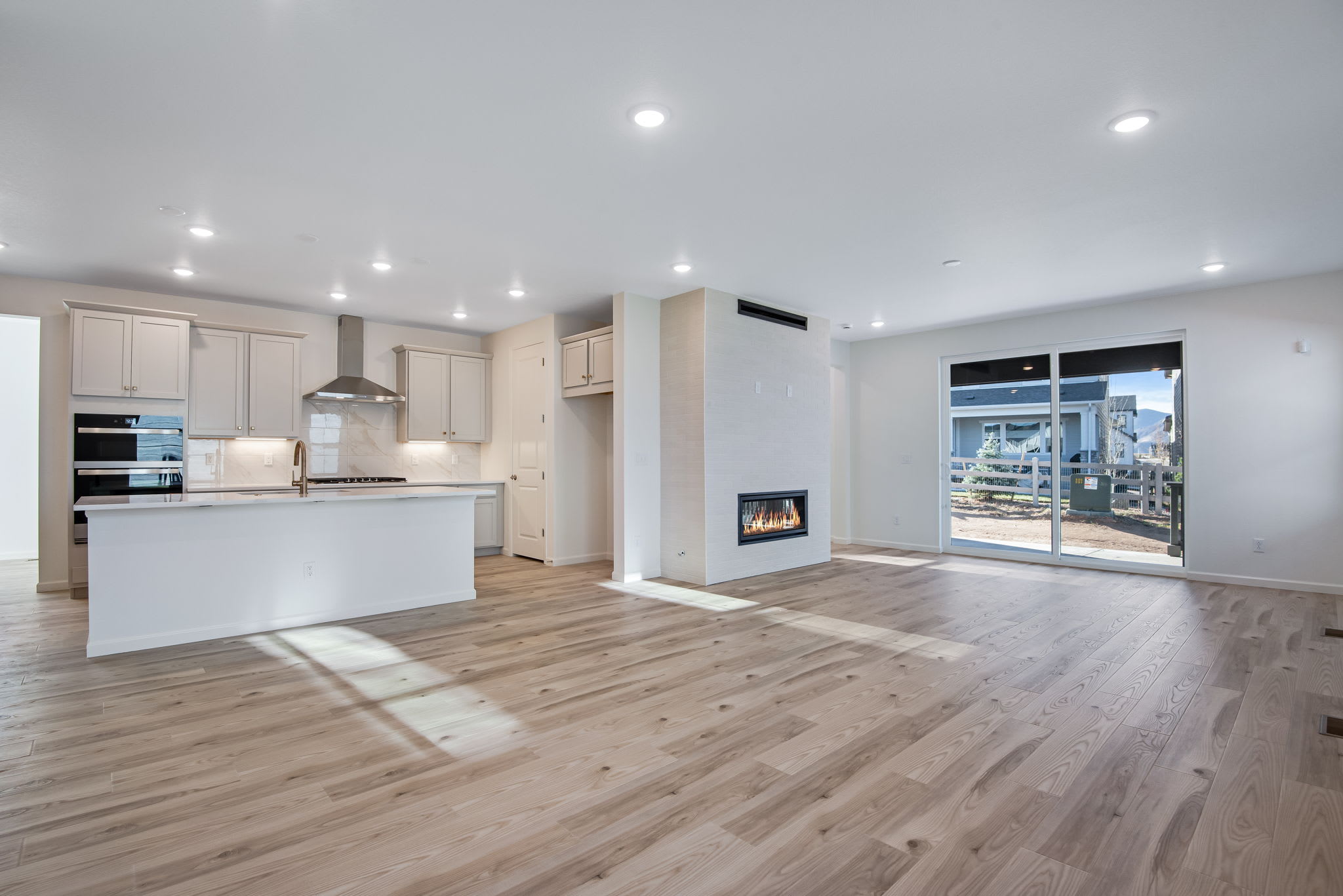 A modern, open-concept kitchen and living room with white cabinets, hardwood floors, and a fireplace visible through a large window overlooking an urban landscape.
