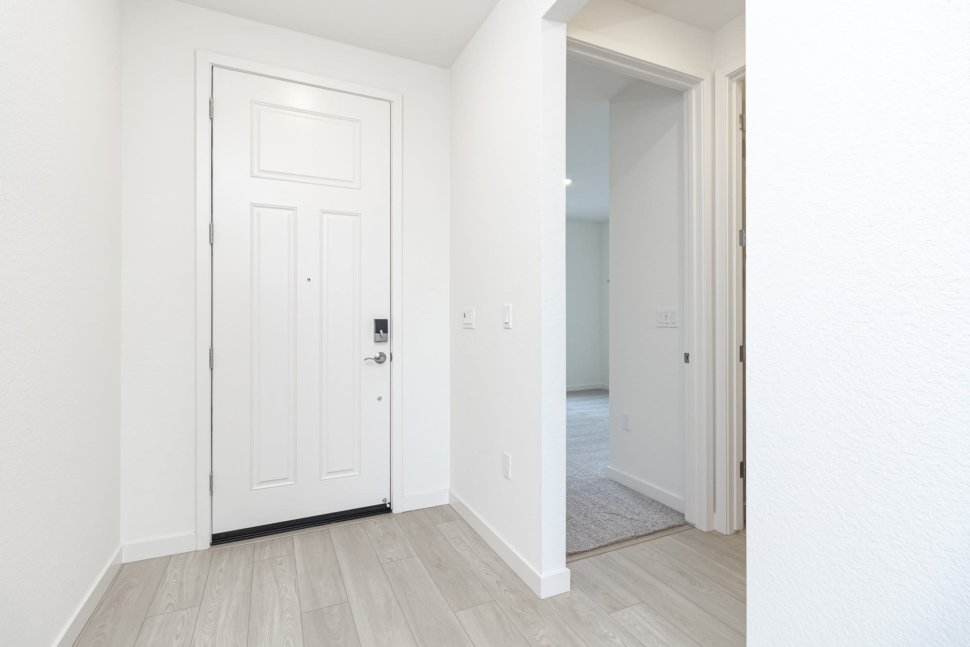 A simple, minimalist entryway with a white door, light-colored wood flooring, and a hallway leading to another room.