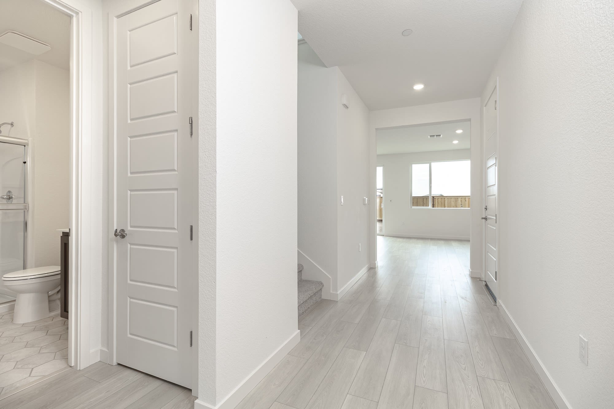 A bright, spacious hallway with white walls, a wooden floor, and a partially visible bathroom door.
