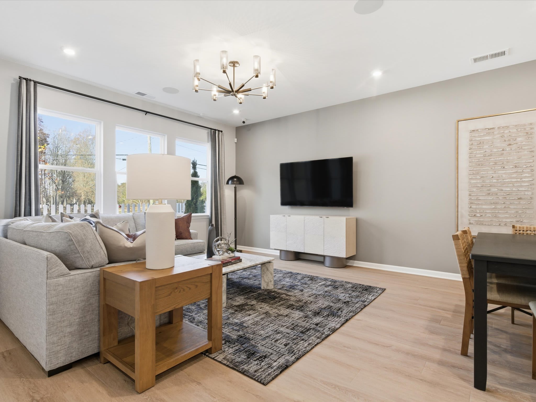 A cozy and modern living room with a gray sofa, a wooden side table, a large TV mounted on the wall, and a chandelier overhead, set against a backdrop of large windows overlooking an outdoor area.