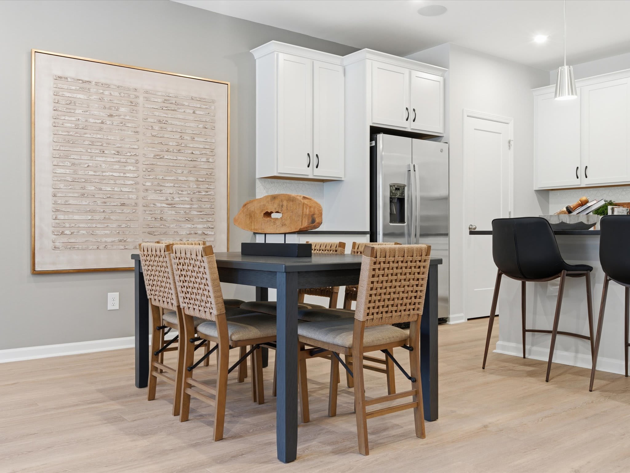 A modern kitchen with white cabinets, a black dining table, and wicker chairs, complemented by a large framed artwork on the wall.