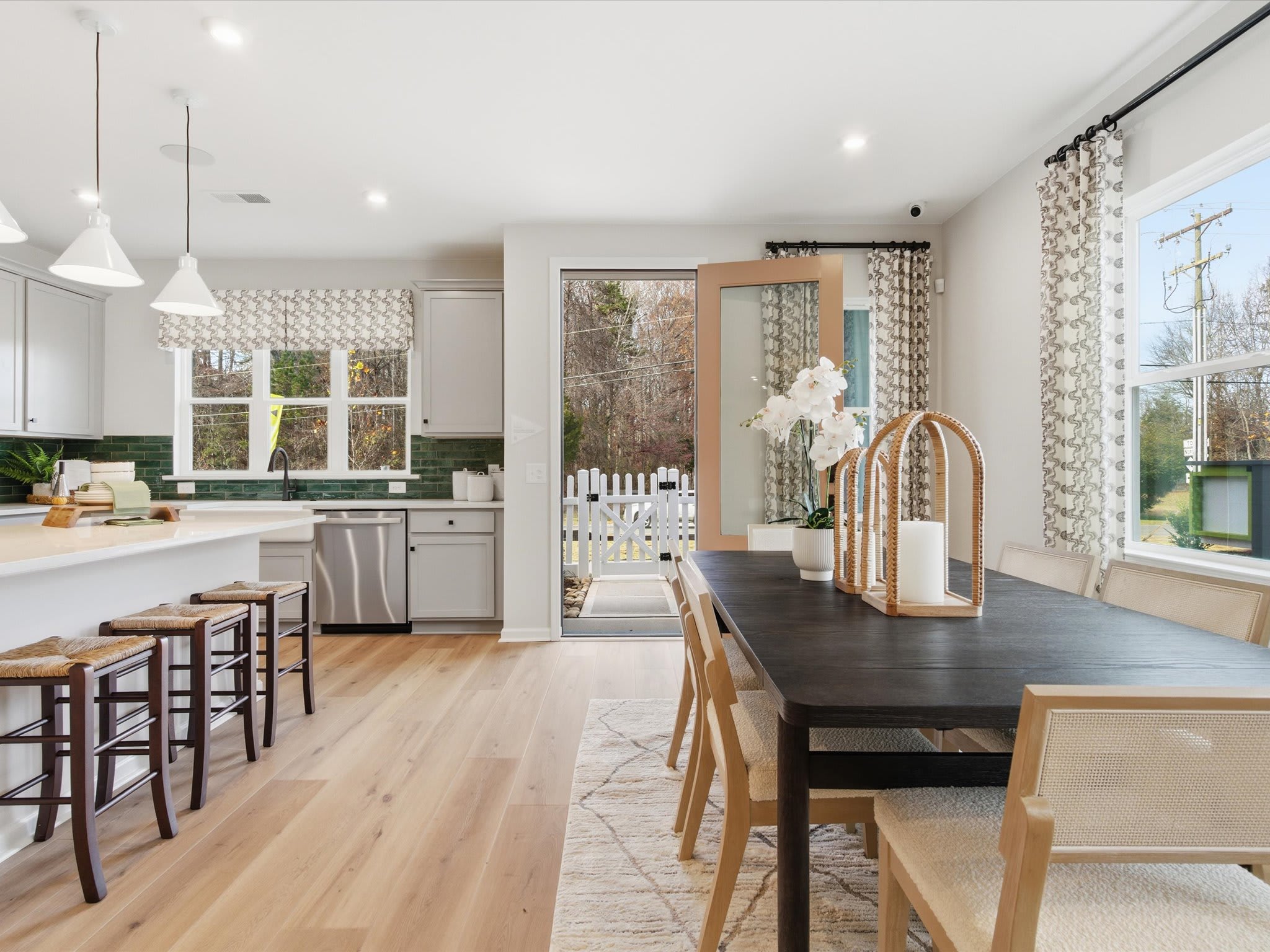 A modern, open-concept kitchen and dining area with wooden floors, white cabinets, and large windows overlooking a wooded outdoor scene.
