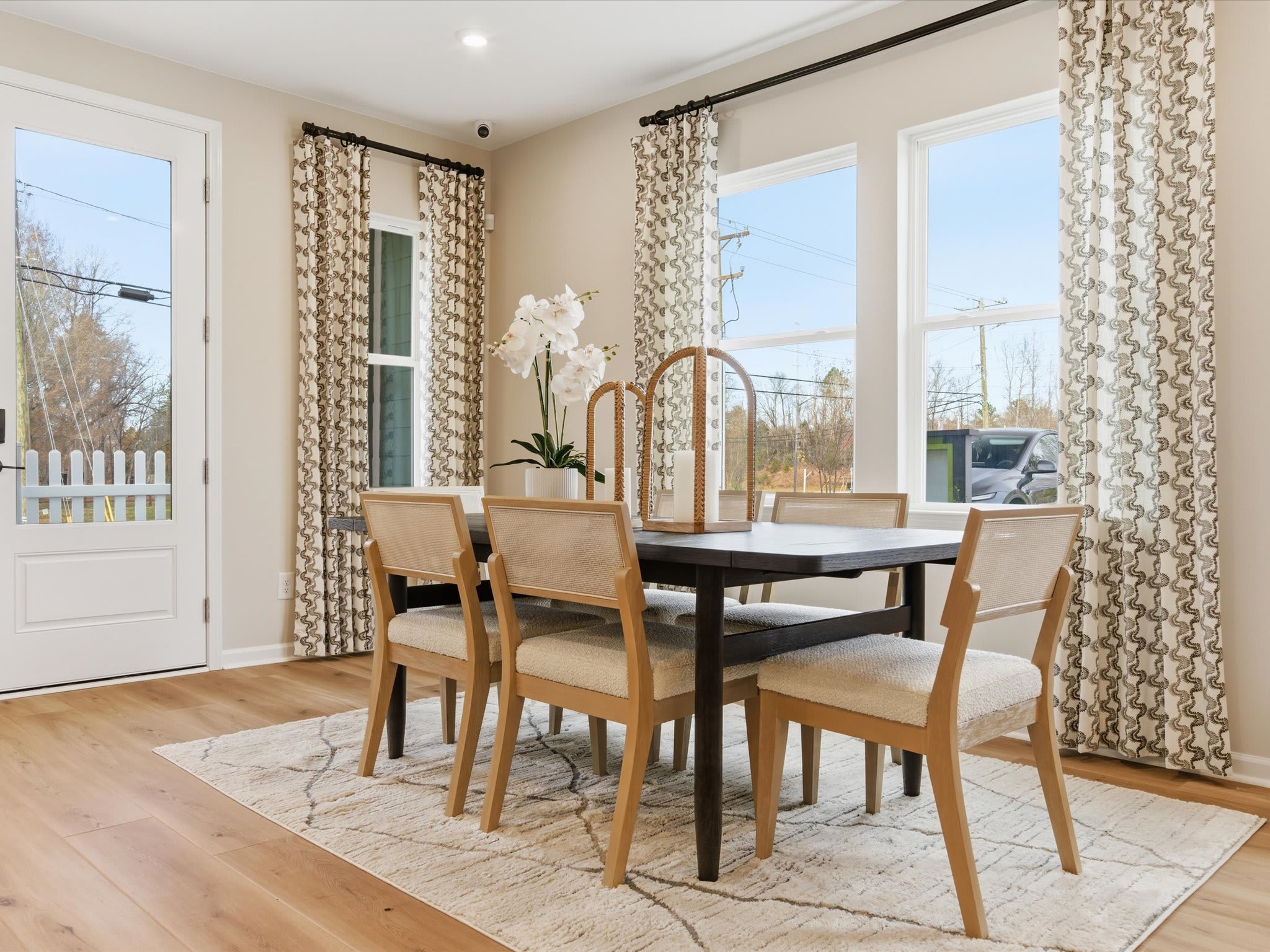 A cozy and well-lit dining room with a wooden table, beige chairs, and patterned curtains, overlooking a scenic outdoor view through large windows.