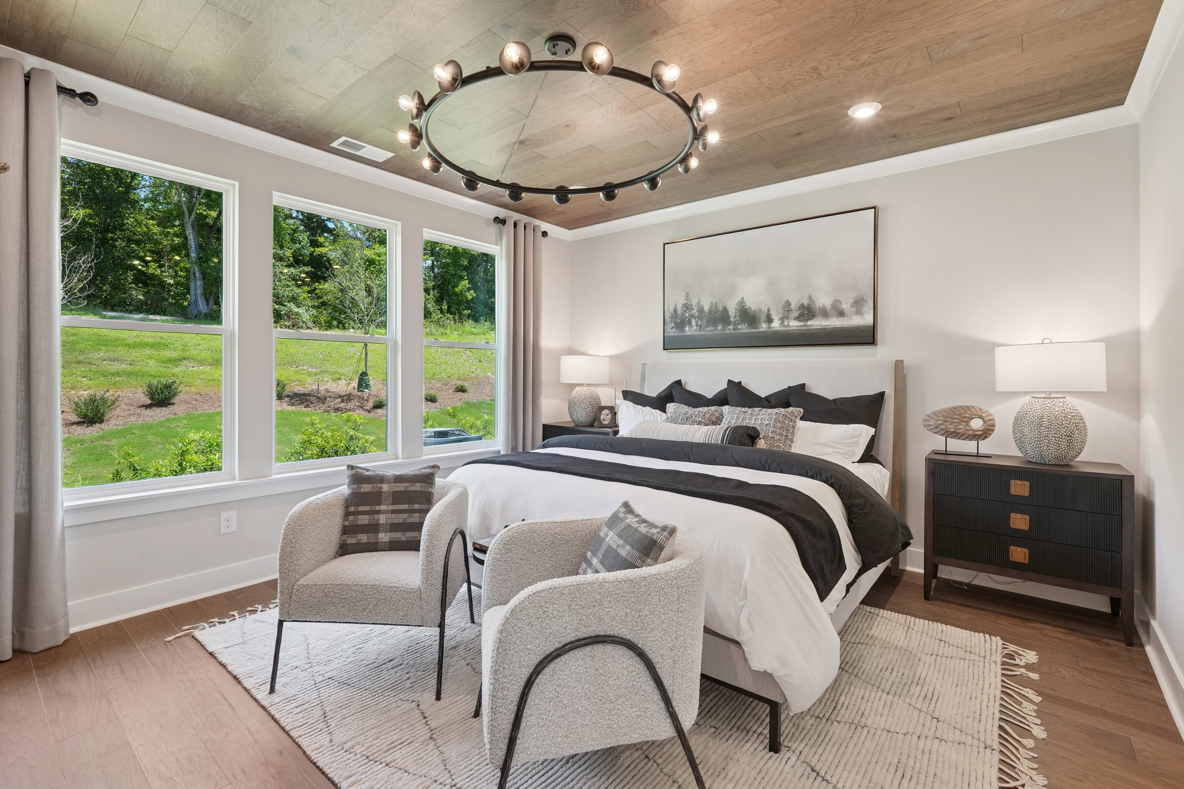 A cozy and inviting bedroom with large windows overlooking a lush, green outdoor landscape, featuring a plush bed, a stylish chandelier, and tasteful decor elements.