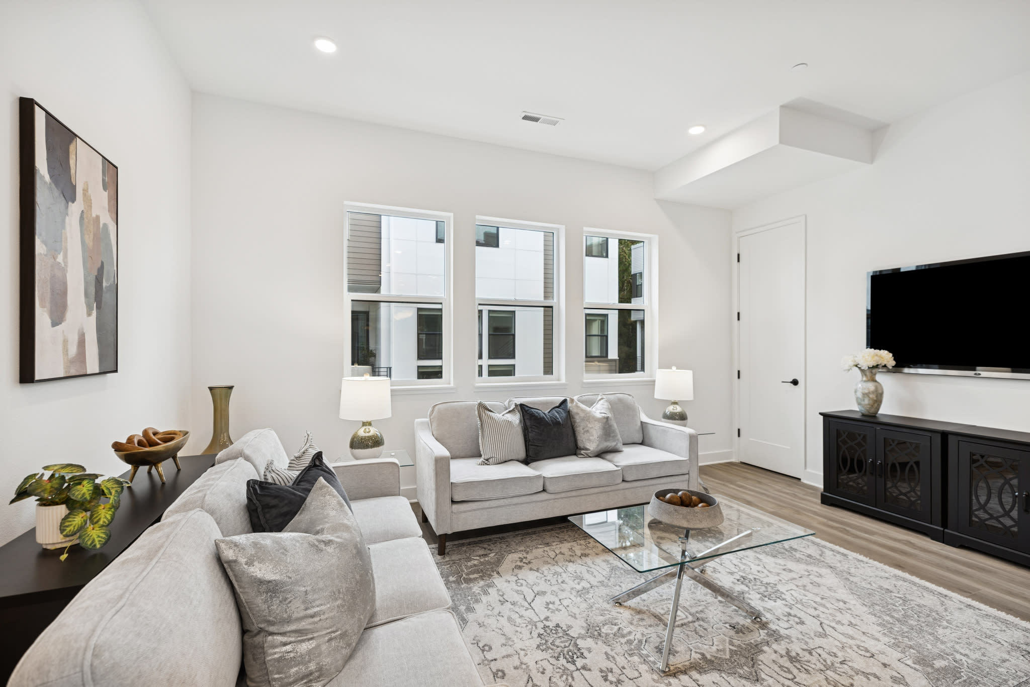 A modern and minimalist living room with a white sofa, glass coffee table, and artwork on the walls, set against a backdrop of large windows and a fireplace.