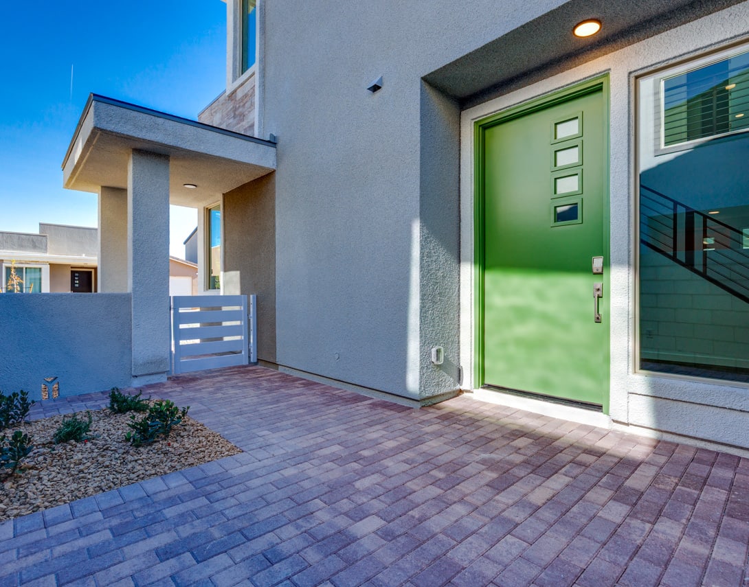 A modern, two-story residential building with a green front door, a paved walkway, and a blue sky in the background.