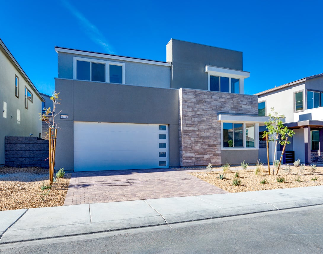 A modern, two-story residential building with a gray exterior, large windows, and a stone accent wall stands in the foreground, set against a clear blue sky in the background.