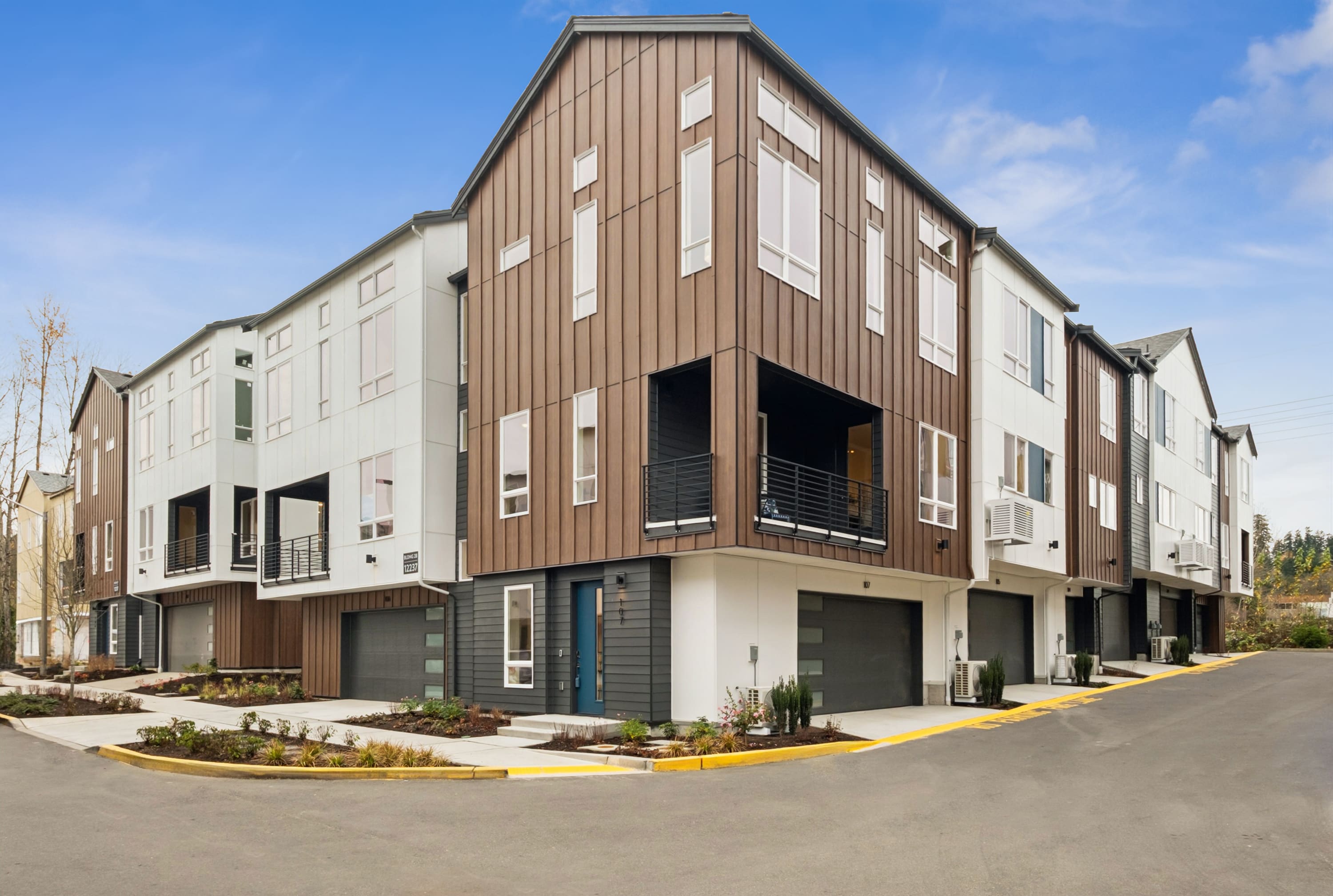 A modern multi-story residential building with a mix of wood and concrete exterior, surrounded by a paved driveway and landscaping in the foreground.