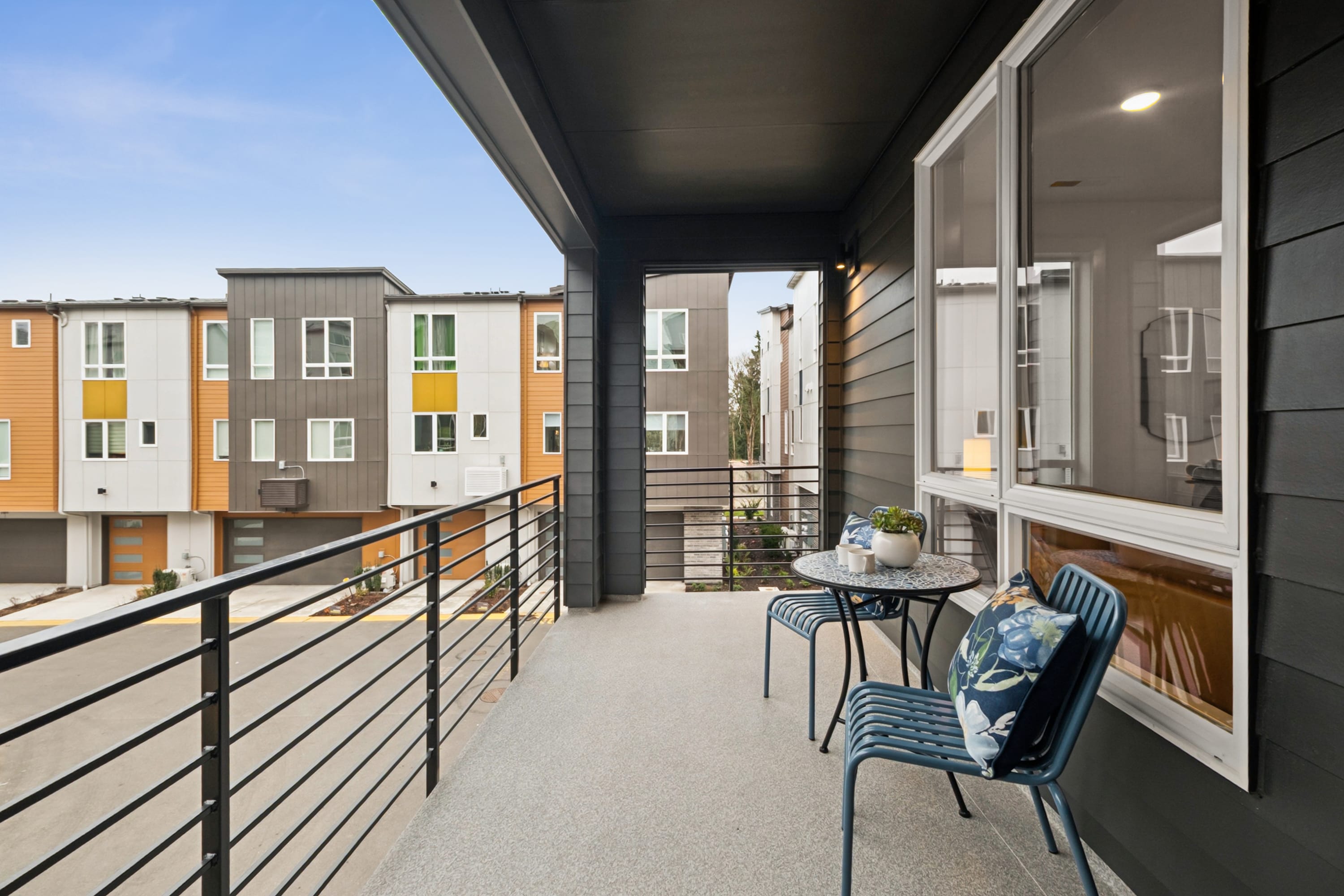 A balcony with a metal railing overlooks a residential neighborhood with colorful, modern townhouses in the background.