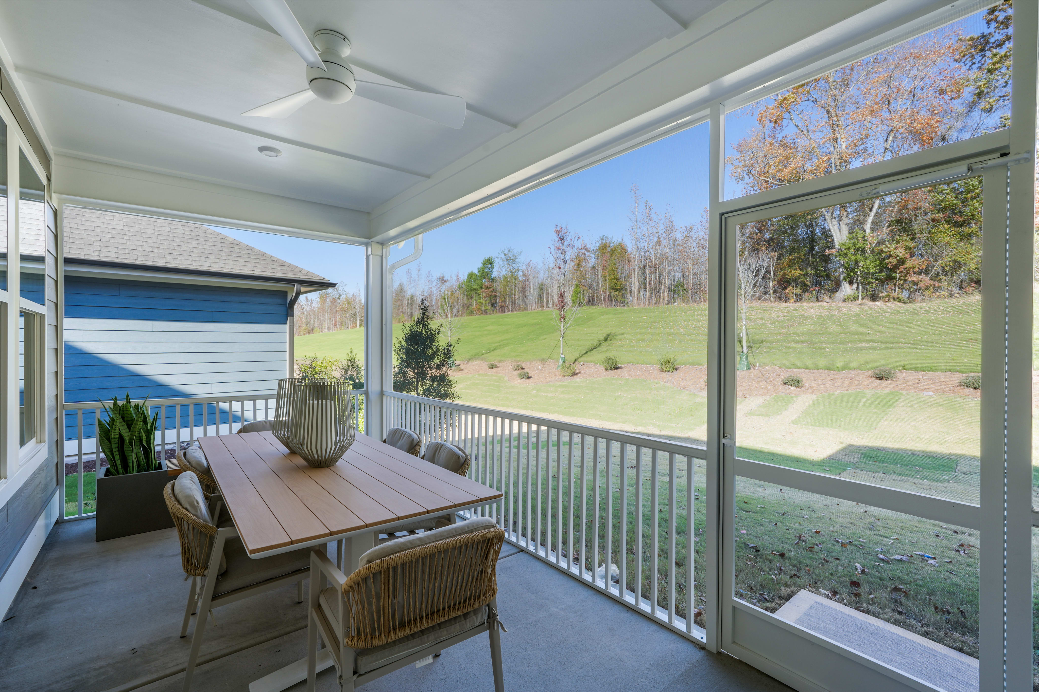 A cozy porch with a wooden table and wicker chairs overlooking a grassy field surrounded by trees in the background.
