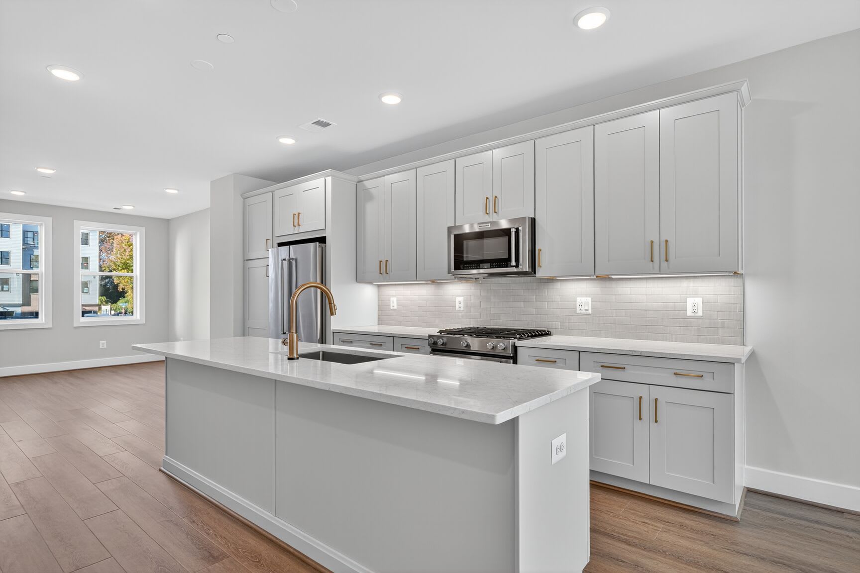 A modern, well-lit kitchen with white cabinets, a central island, and hardwood floors, set against a backdrop of large windows providing natural light.