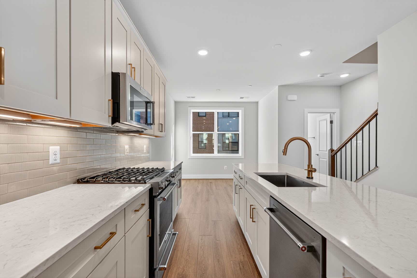 A modern, well-lit kitchen with white cabinets, stainless steel appliances, and a wooden floor leading to a hallway.