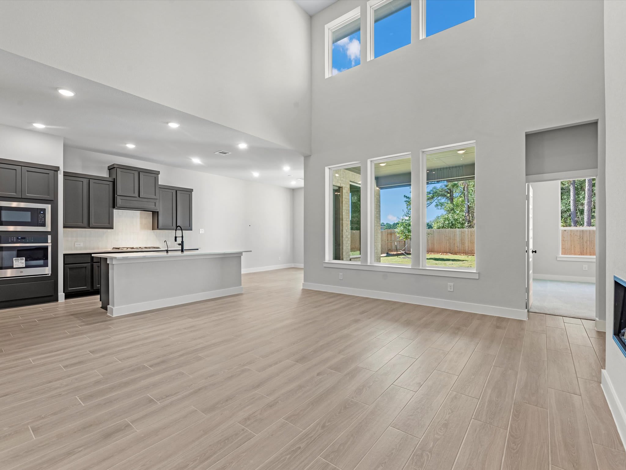 A spacious and modern open-plan living area with a kitchen, hardwood floors, and large windows overlooking a backyard.