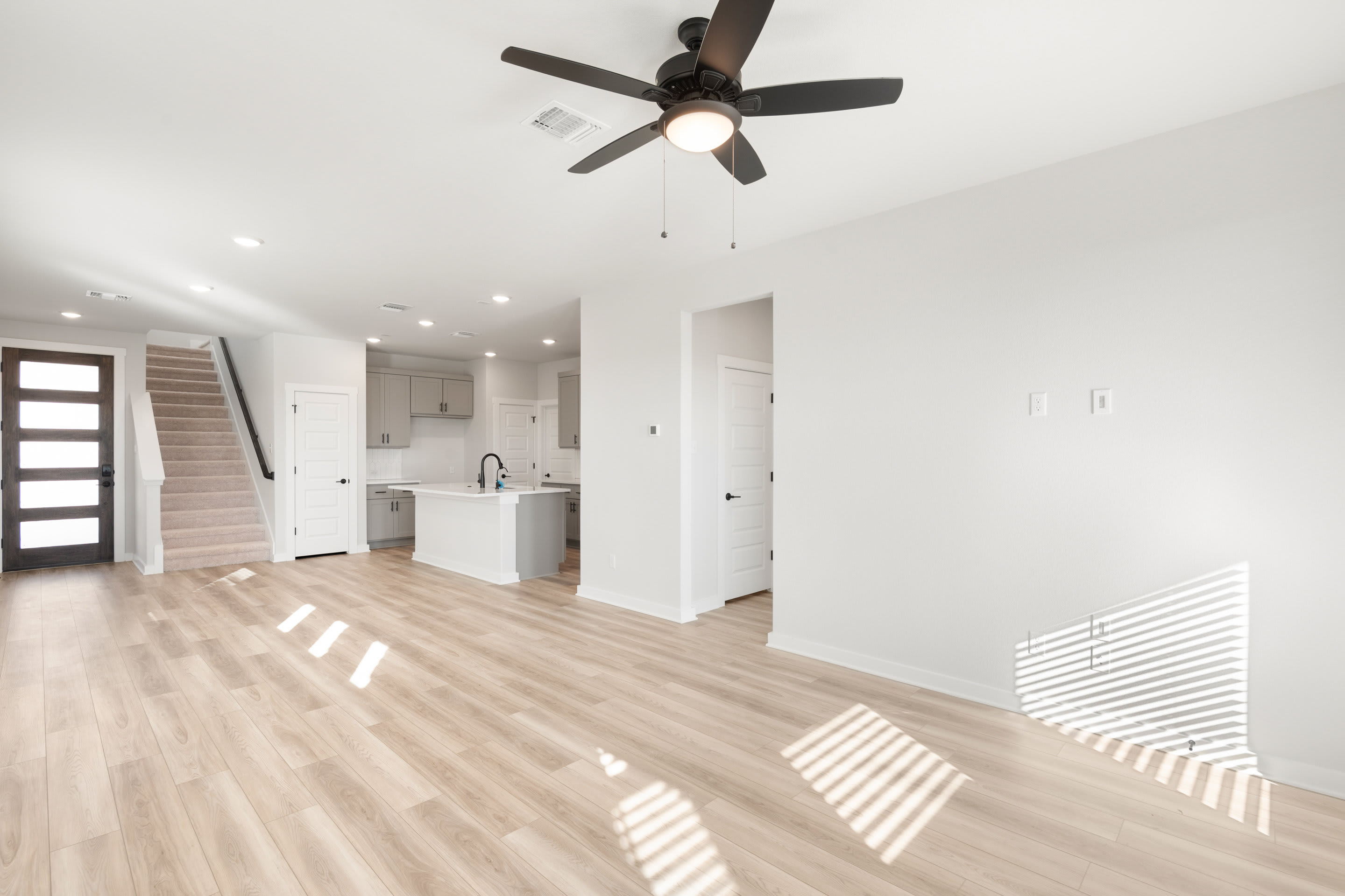 A bright, open-concept living space with a ceiling fan, hardwood floors, and a view of a kitchen area in the background.