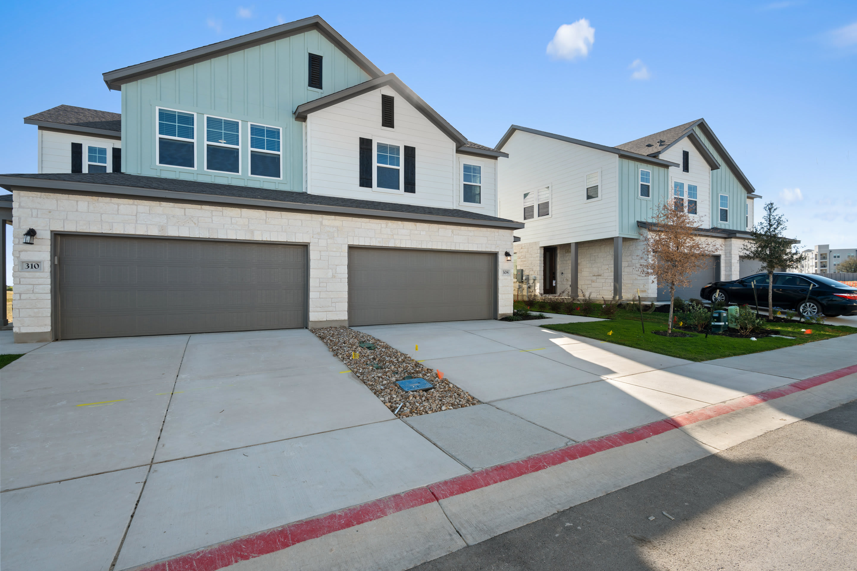 A modern, multi-story residential building with a garage and a paved driveway in the foreground, set against a clear blue sky with some clouds.