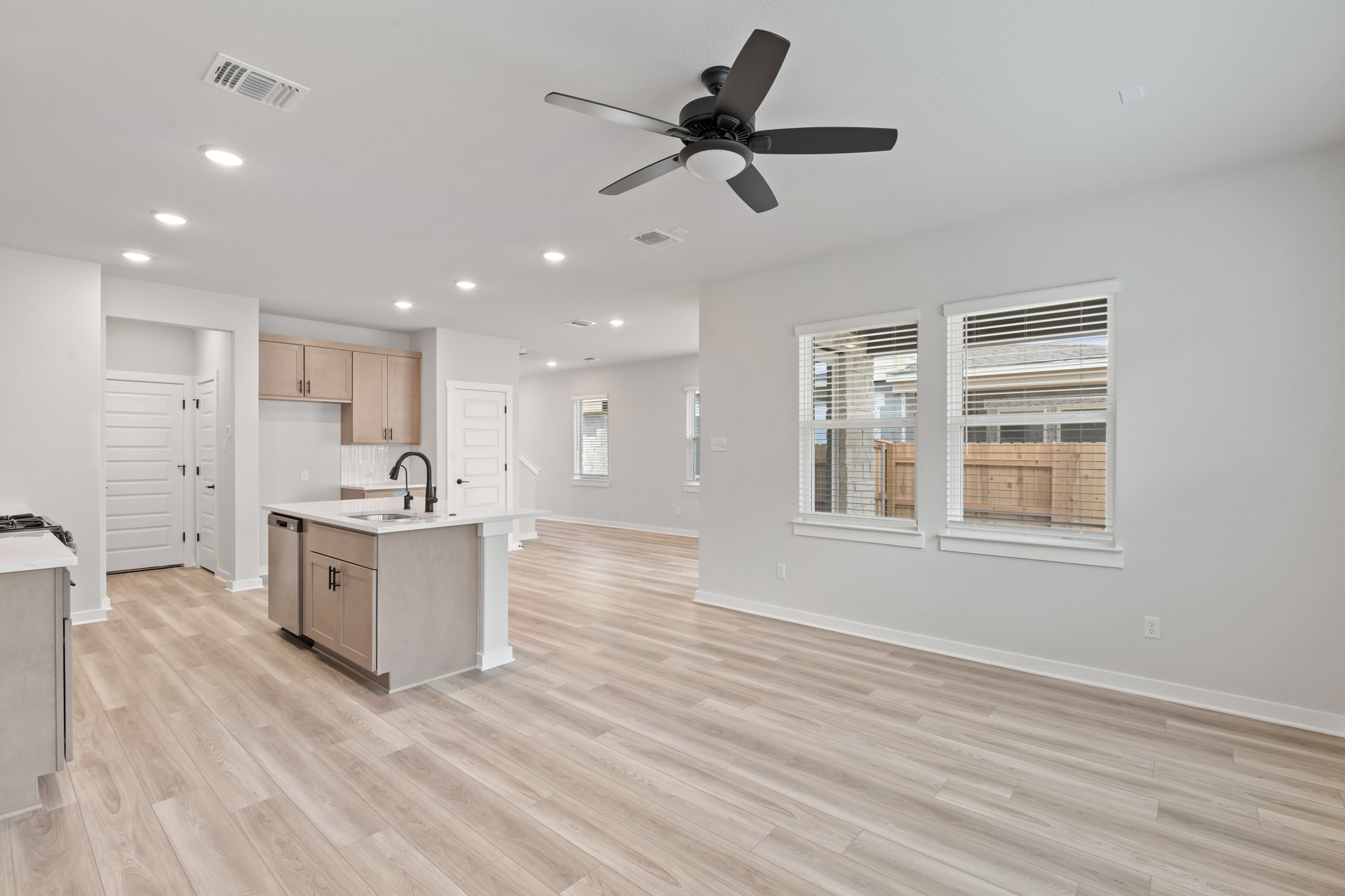 A modern, open-concept kitchen and living space with a ceiling fan, hardwood floors, and large windows providing natural light.
