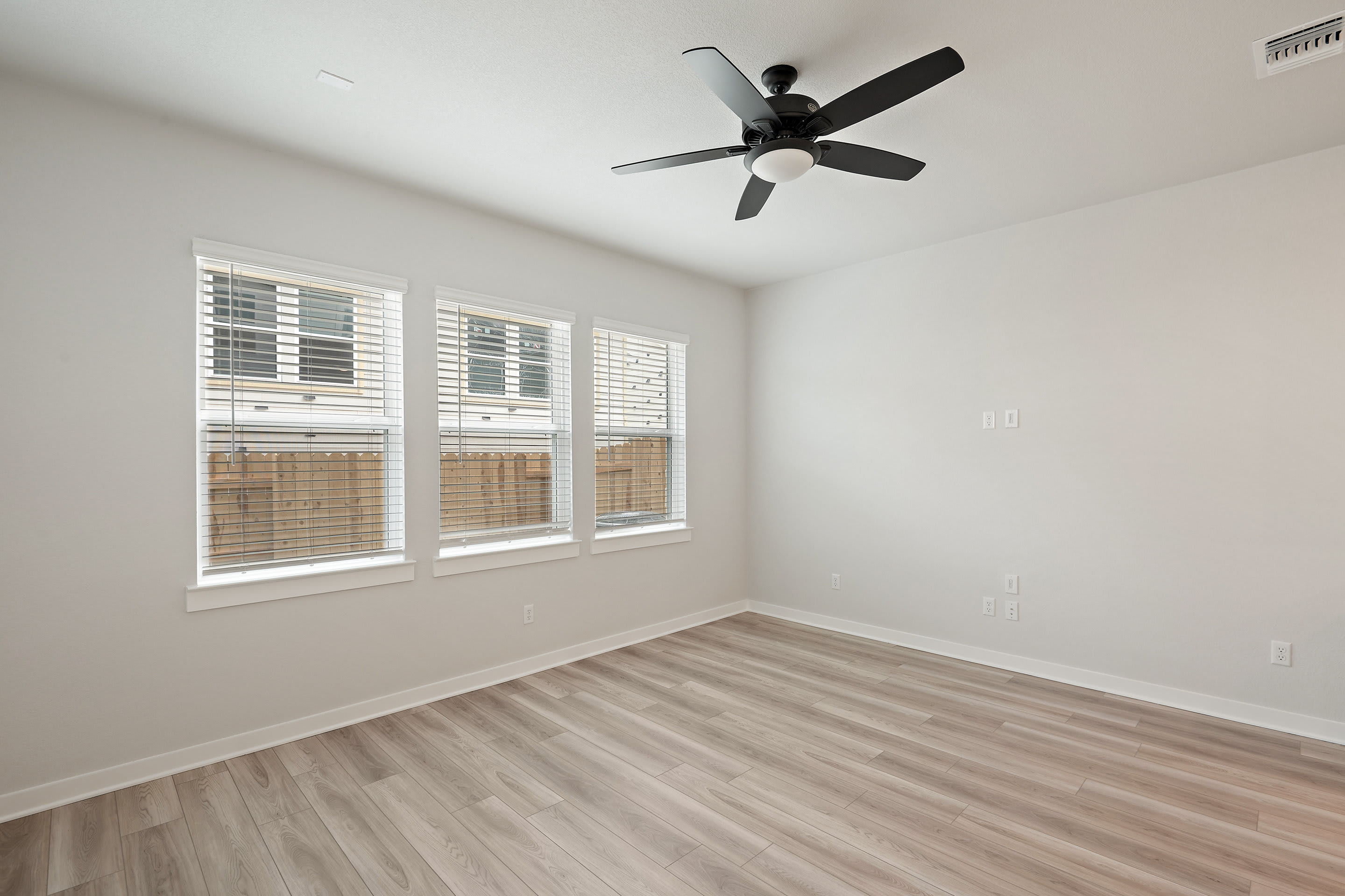 A spacious, well-lit room with a ceiling fan, wooden blinds, and a hardwood floor.