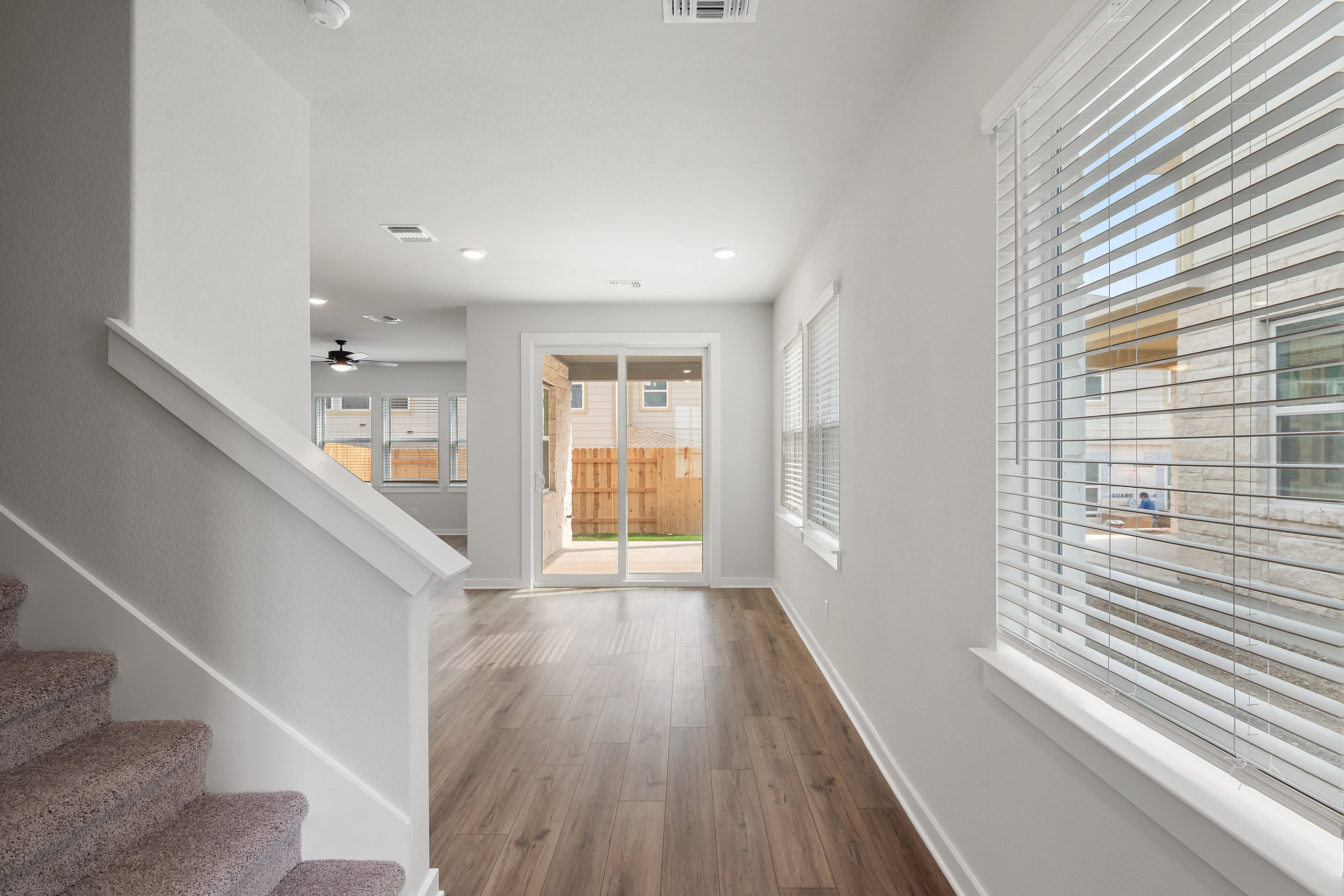 A bright, spacious hallway with wooden floors, white walls, and a set of stairs leading to the upper level. The hallway is well-lit, with a glass door visible in the background, allowing natural light to flood the space.
