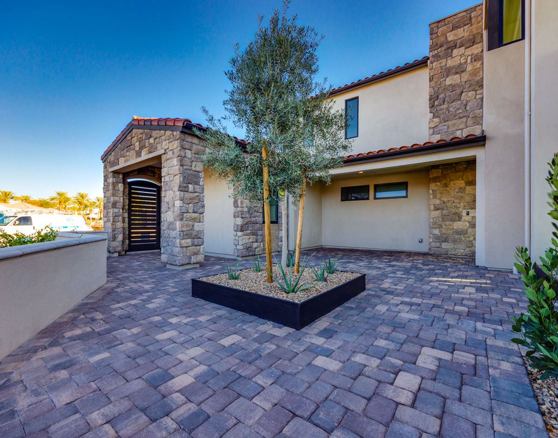 A well-designed outdoor patio area with a stone-clad building, a paved brick floor, and a potted tree in the foreground, set against a clear blue sky.
