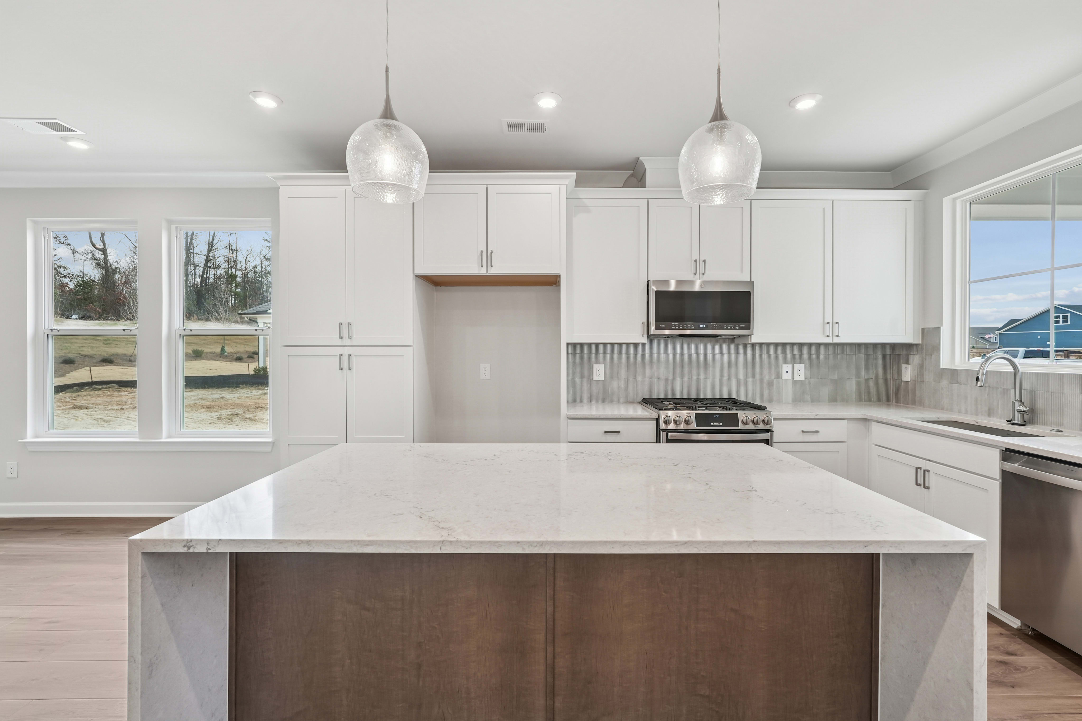 A modern, bright kitchen with white cabinets, a large island, and pendant lighting, set against a backdrop of windows overlooking a scenic outdoor landscape.