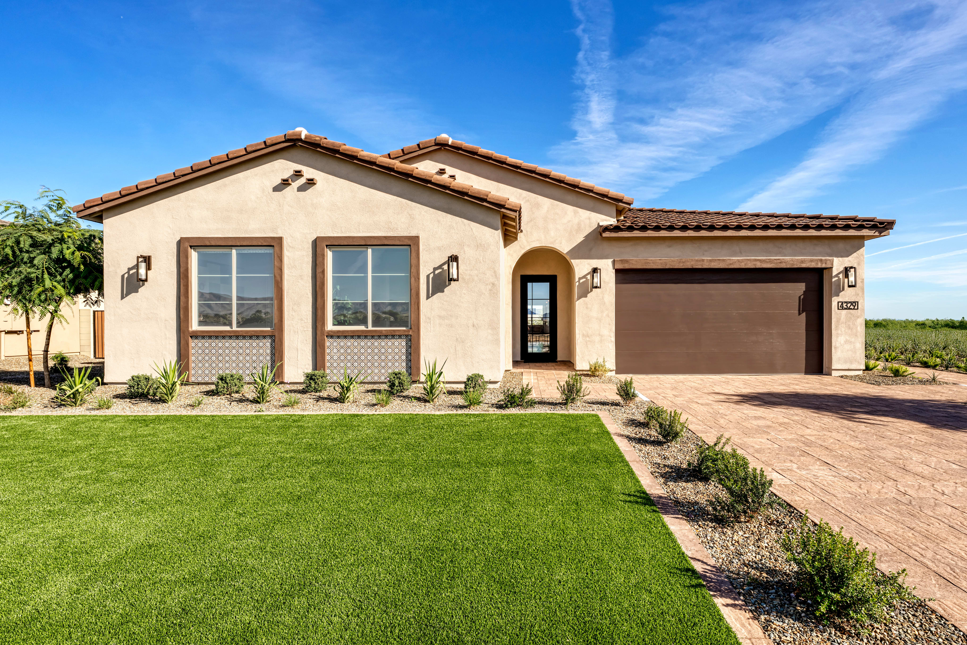 A well-manicured lawn leads to a two-story Mediterranean-style house with a tiled roof, surrounded by lush greenery and a clear blue sky.