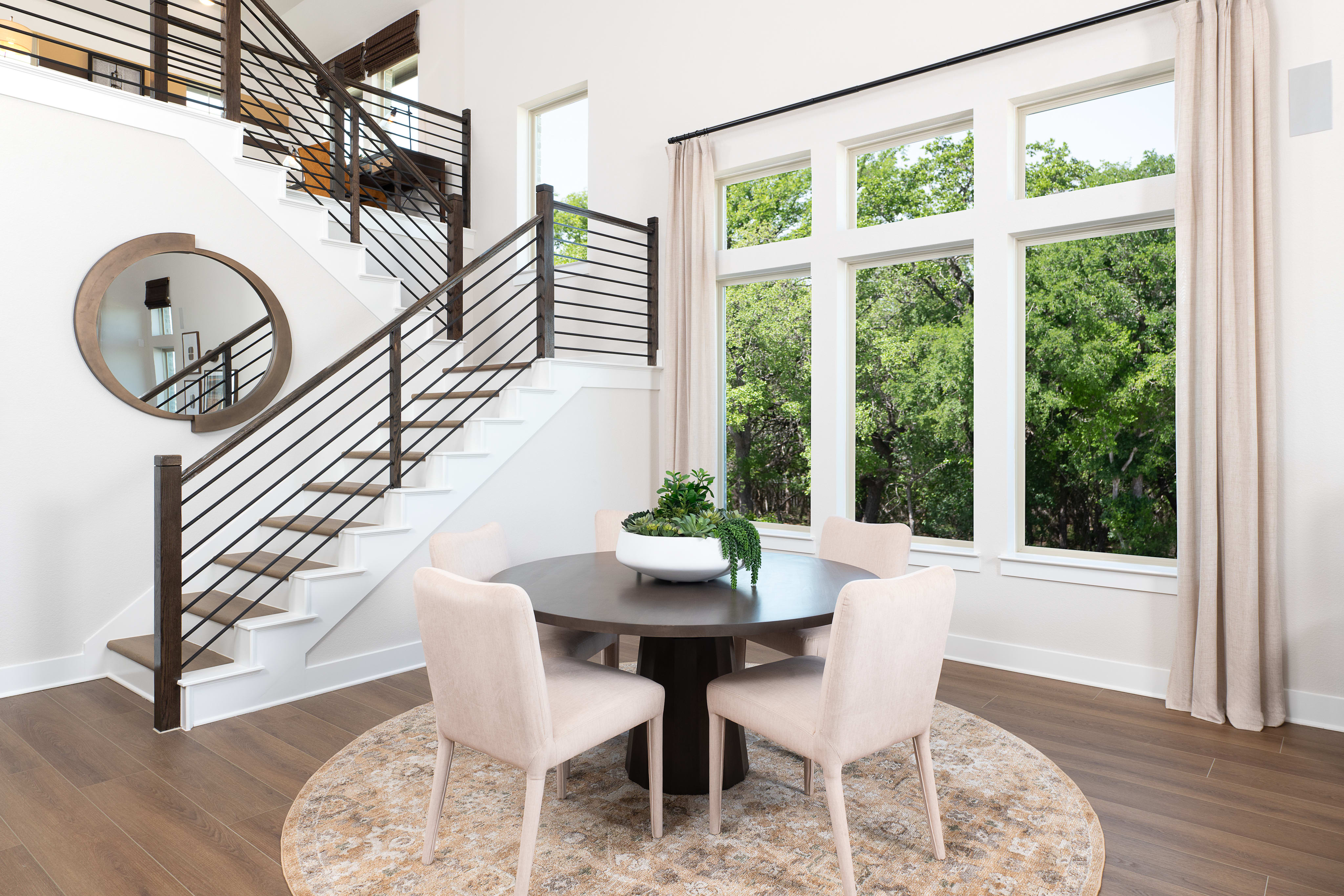 A modern and airy dining area with a round wooden table and white chairs, surrounded by large windows overlooking a lush, green outdoor landscape.
