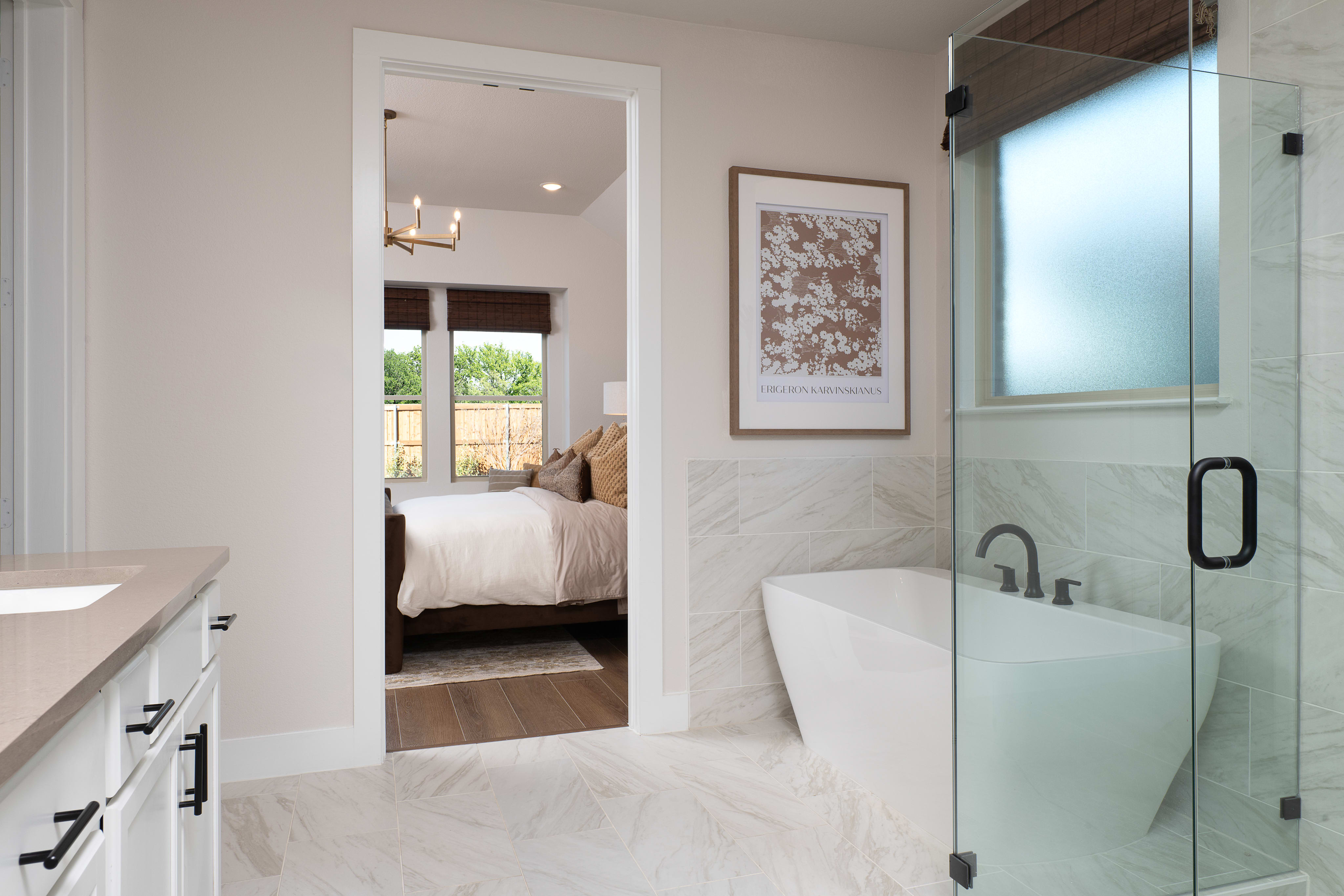 The image depicts a modern and minimalist bathroom with a glass shower enclosure, a white vanity, and a framed artwork on the wall. In the background, a bedroom can be seen through an open doorway.