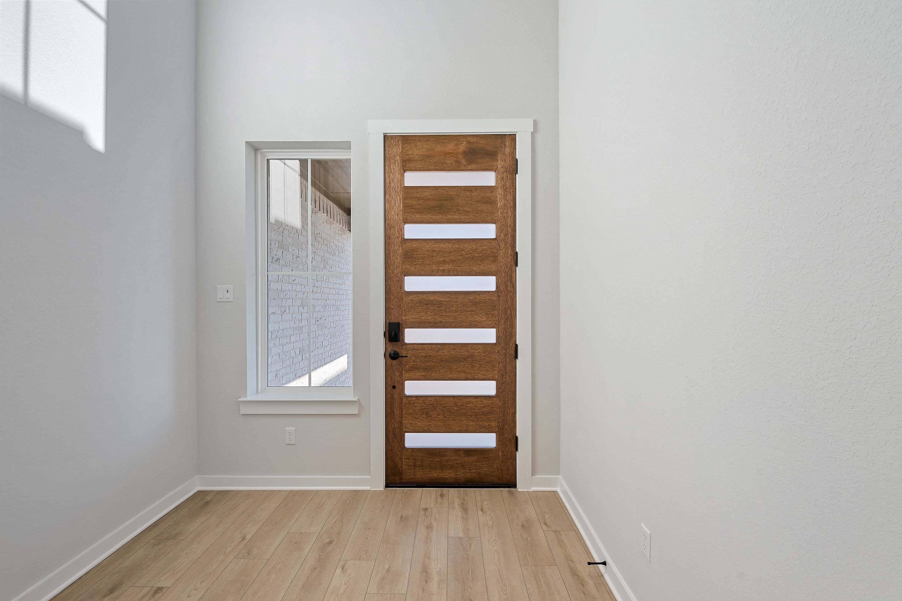 A wooden door with horizontal panels stands in a bright, minimalist room with a window and hardwood flooring.