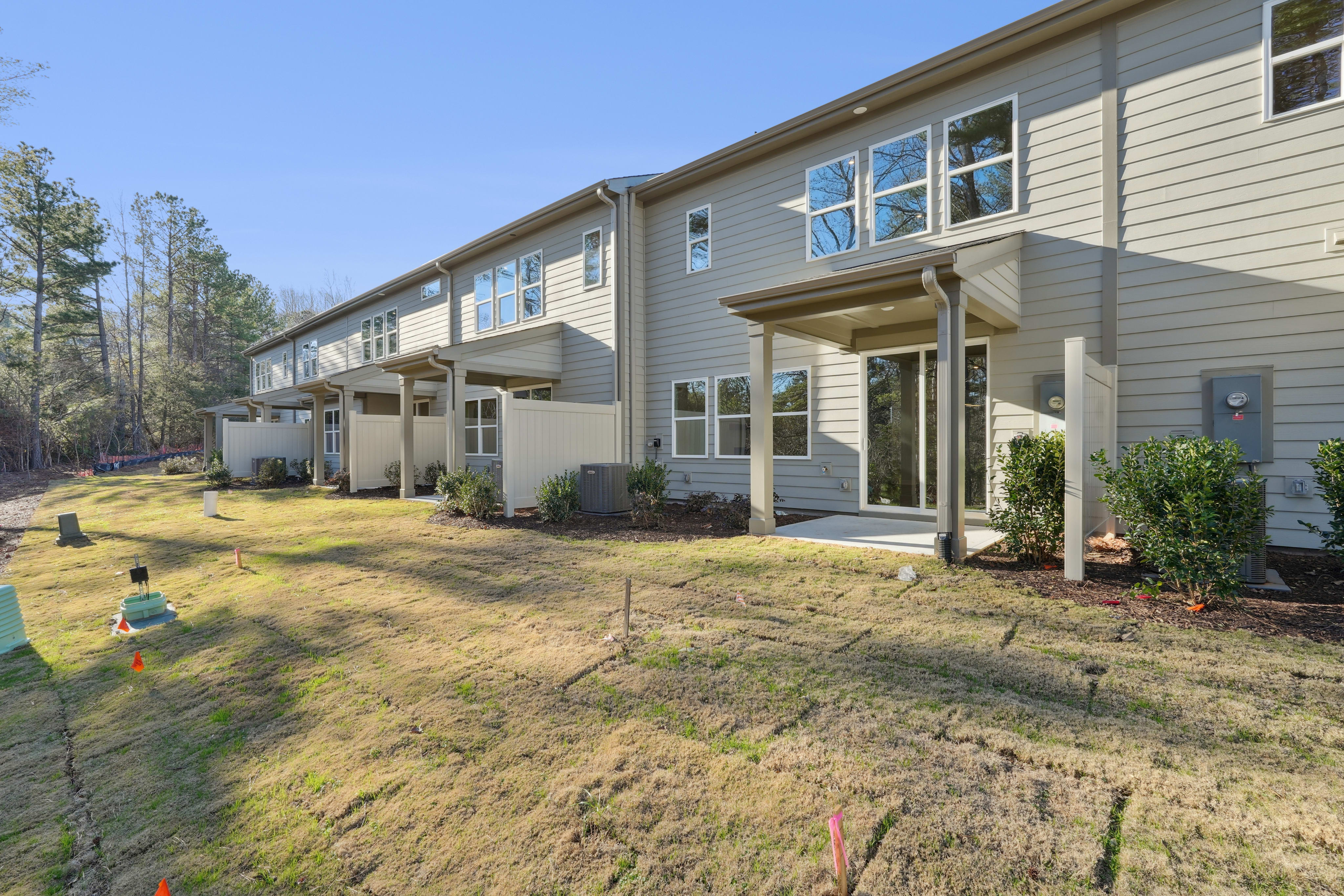 A row of beige-colored townhouses with landscaped yards and a grassy area in the foreground, set against a backdrop of pine trees.