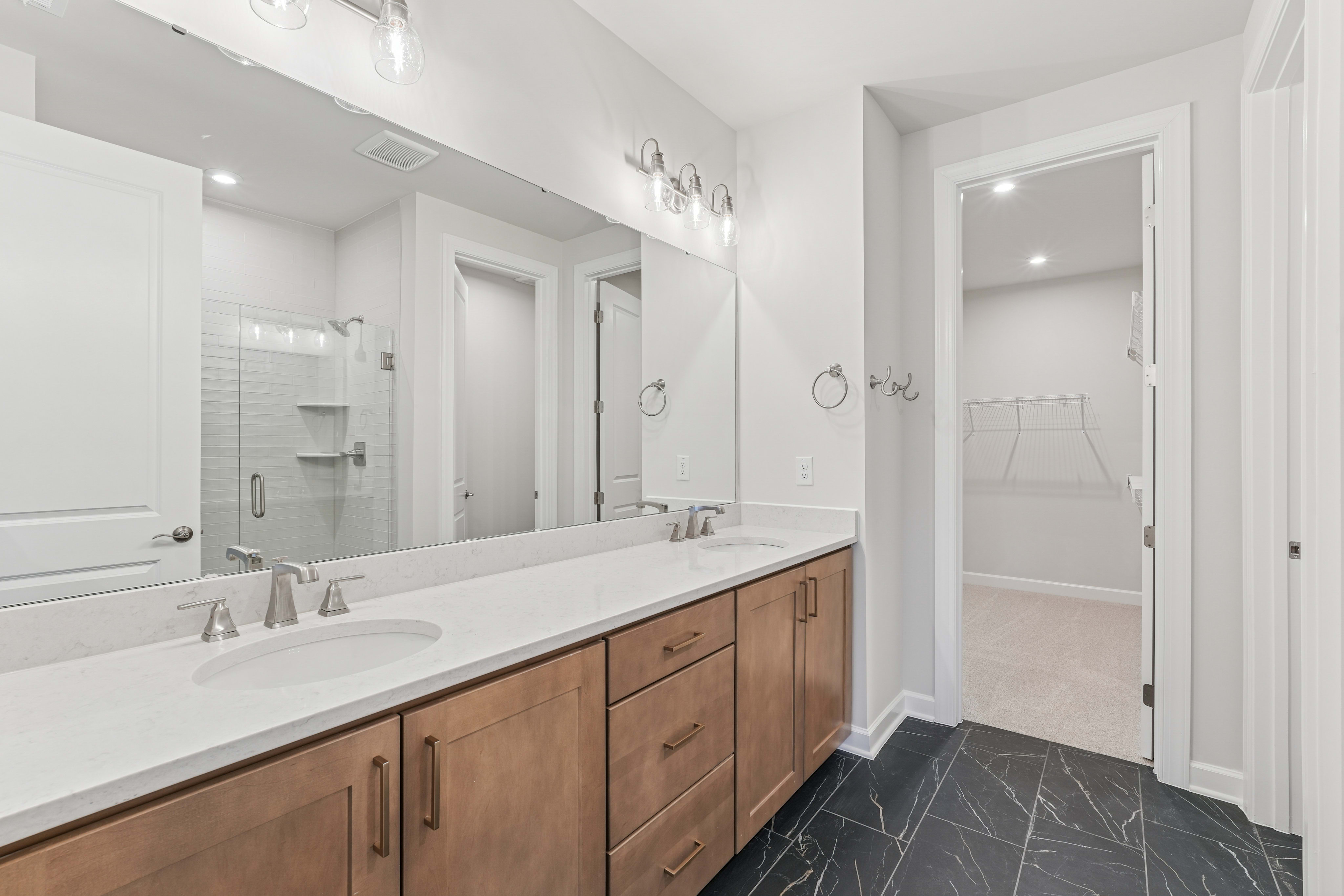 A modern and spacious bathroom with a double vanity, white countertops, and a tiled floor. The room features recessed lighting and a glass-enclosed shower area.