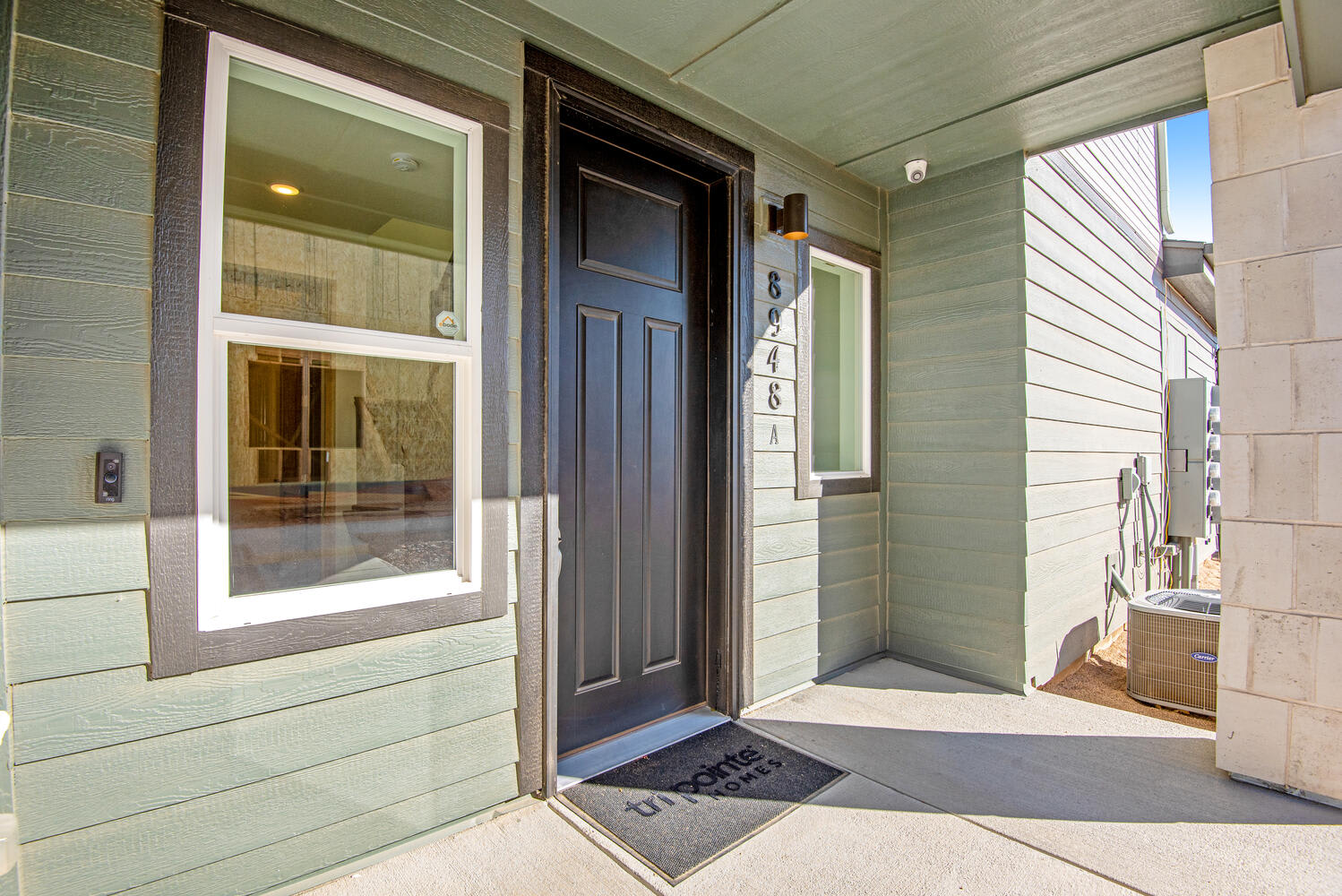 The image shows a well-lit entryway with a dark wooden door, a glass-paneled door, and a gray welcome mat on the porch. The walls are painted a light green color, and there are some decorative elements visible in the background.