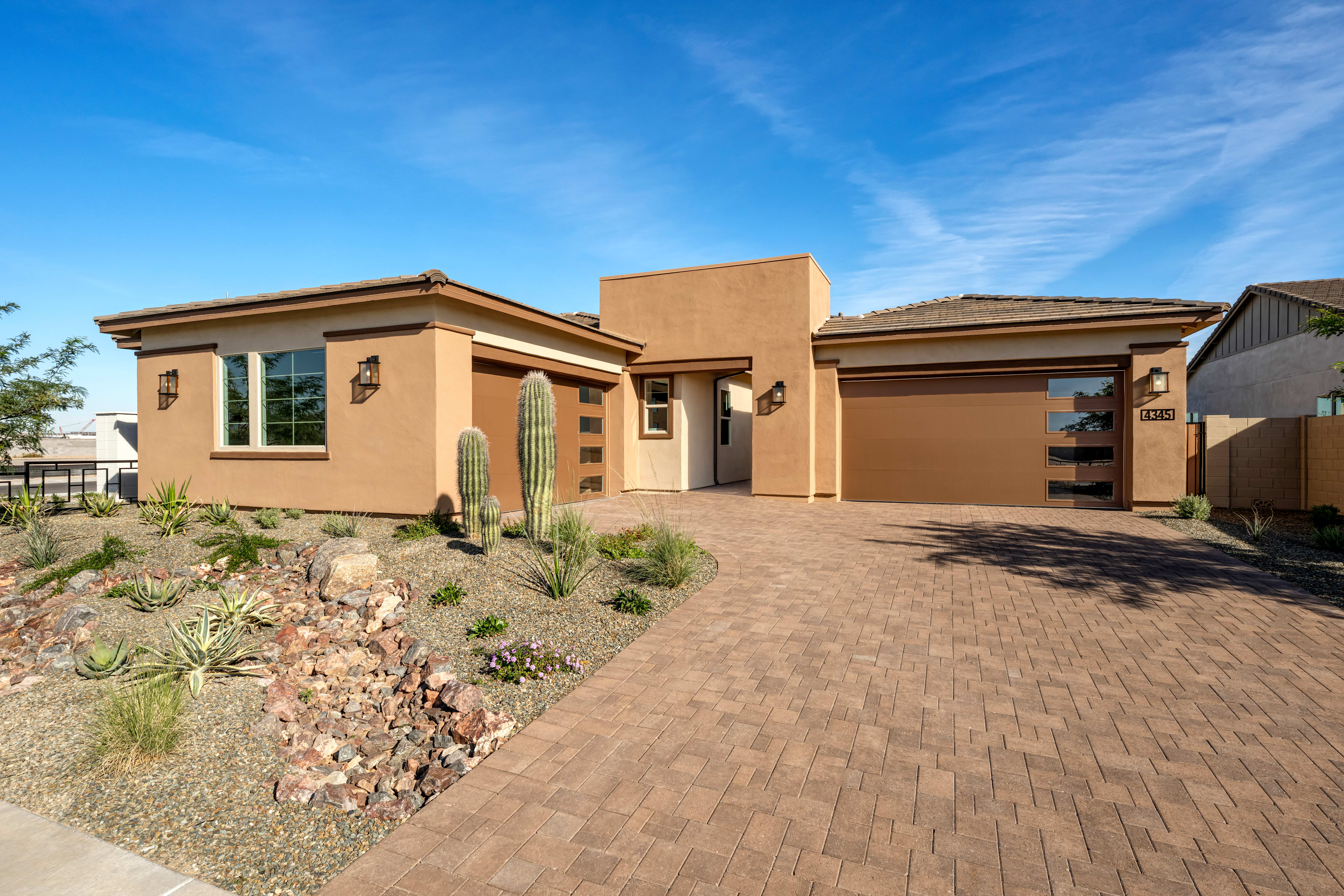A single-story, stucco-clad house with a tiled roof sits on a paved driveway surrounded by desert landscaping, against a backdrop of a clear blue sky with wispy clouds.