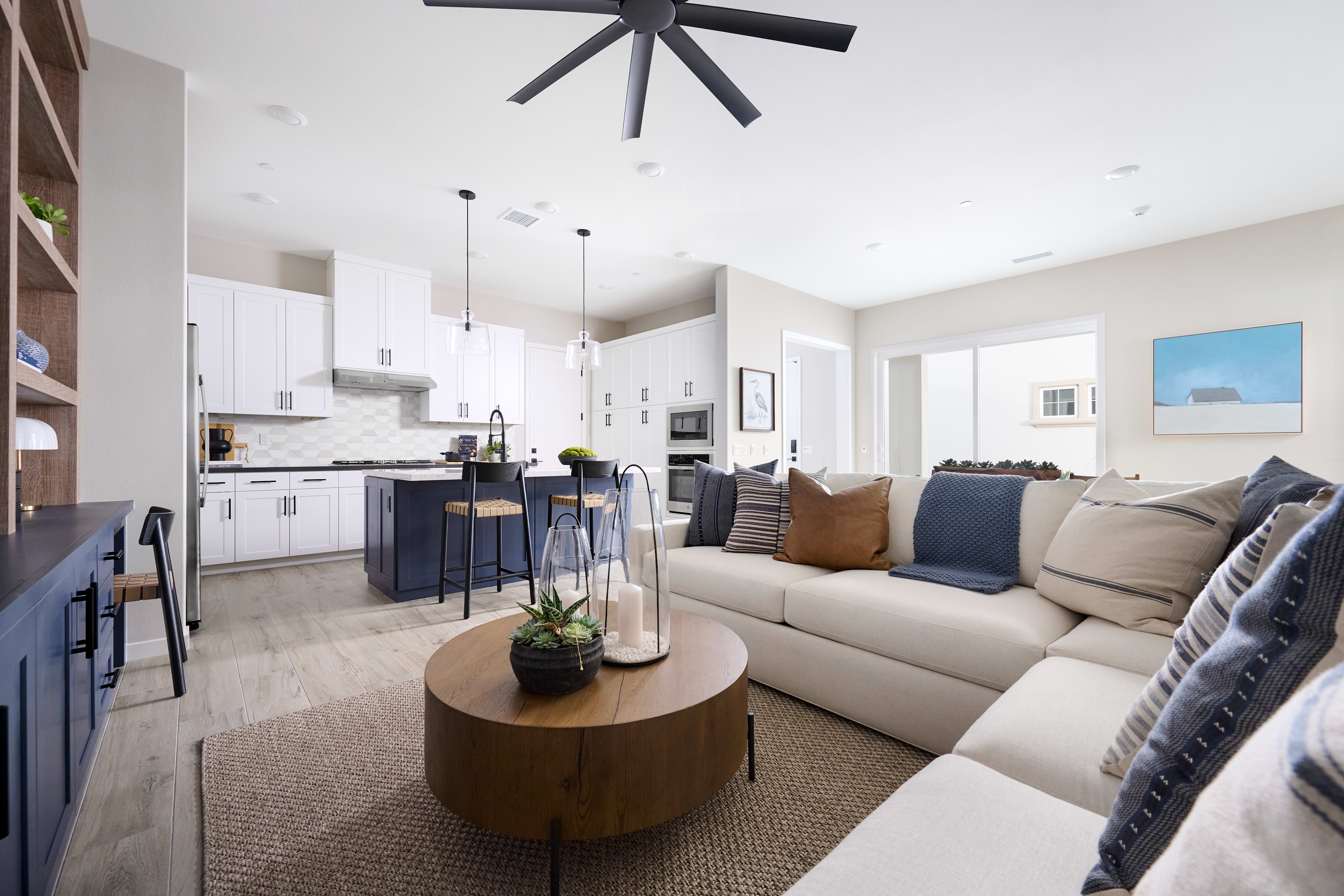 A modern and cozy open-concept living space with a white sectional sofa, a round wooden coffee table, and a large ceiling fan in the background.