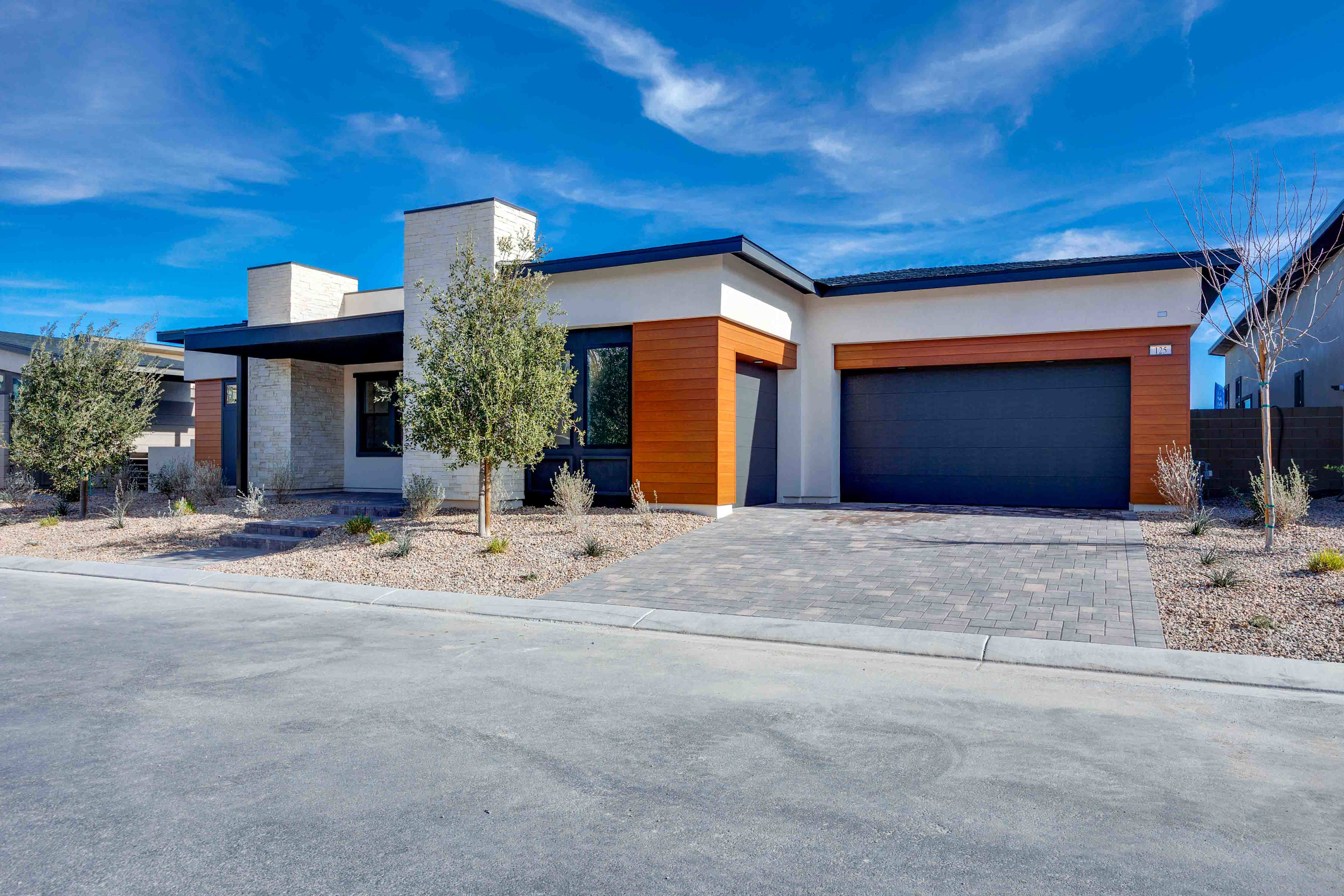 A modern, single-story house with a garage, surrounded by a paved driveway and landscaped with desert vegetation against a backdrop of a clear blue sky with wispy clouds.