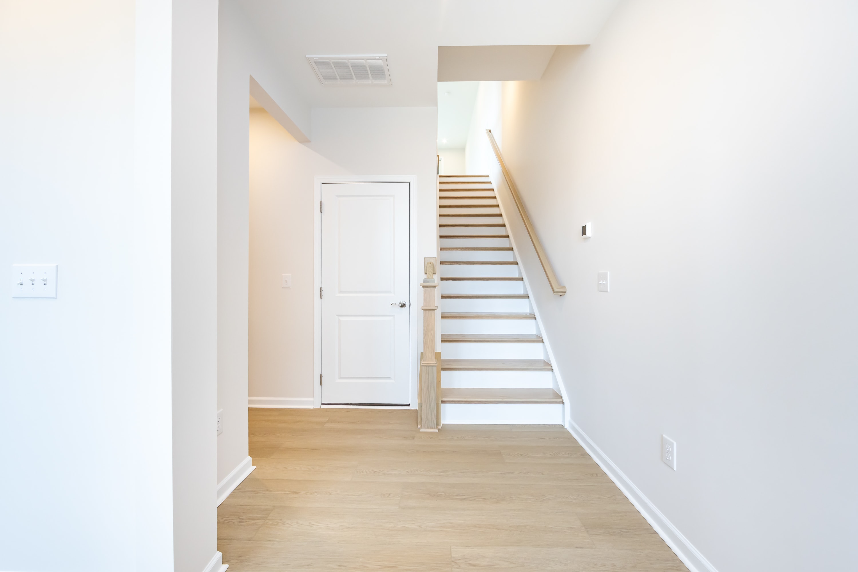 A bright and airy hallway with a wooden staircase leading upstairs, and a white door visible in the background.