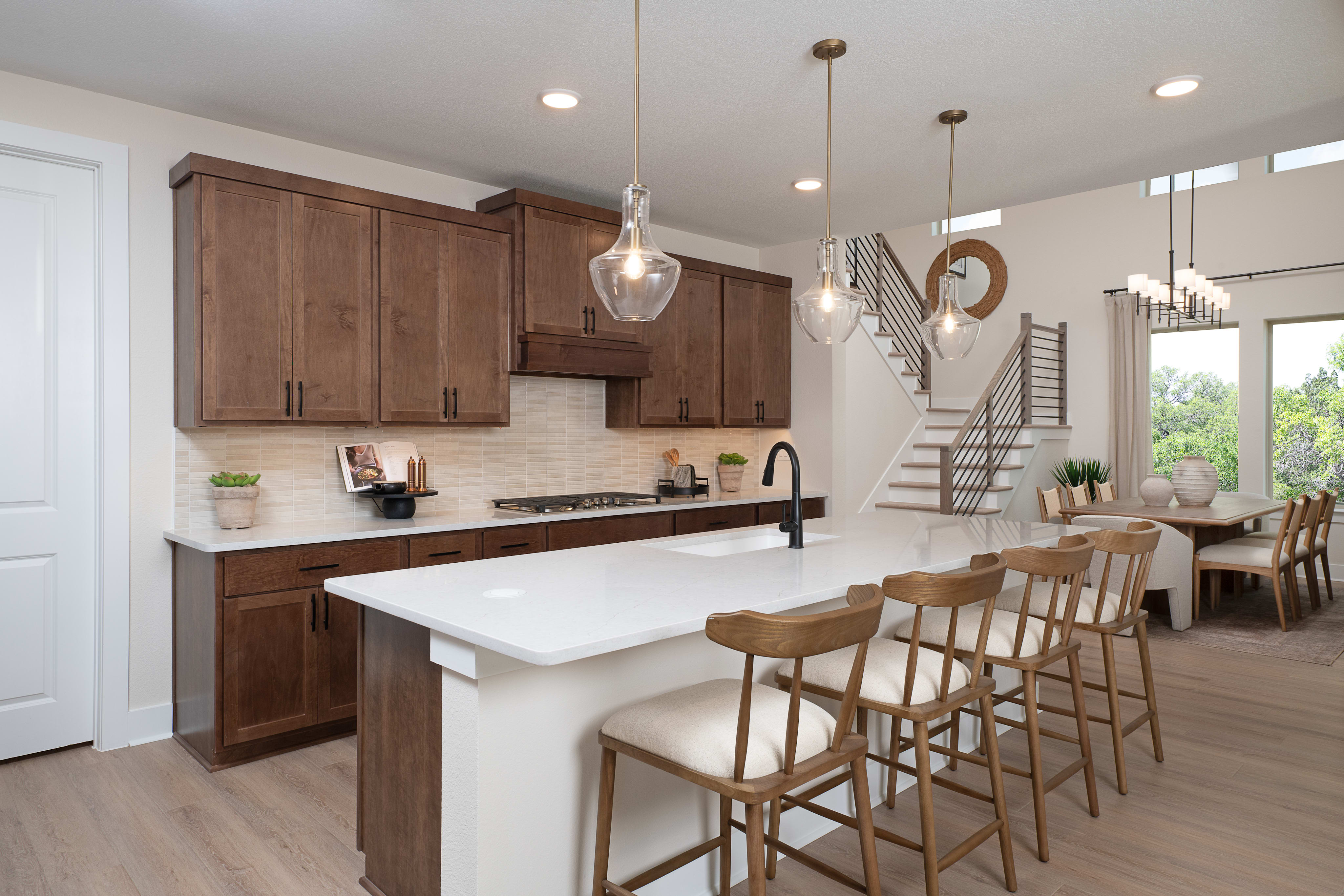 A modern, open-concept kitchen with wooden cabinets, a white countertop, and pendant lighting, leading into a dining area with a wooden table and chairs.