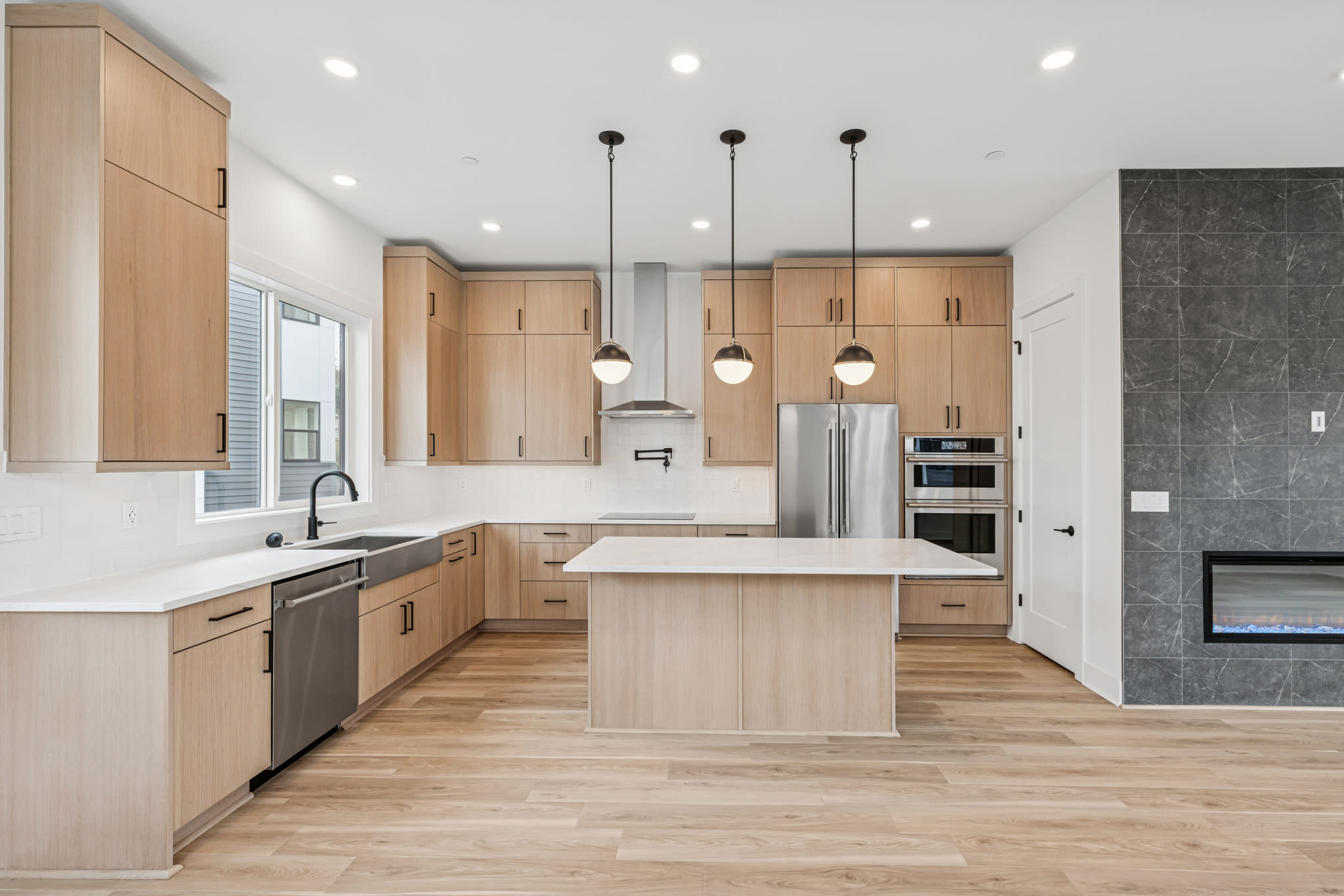 A modern, open-concept kitchen with light wood cabinetry, stainless steel appliances, and a central island with a white countertop.