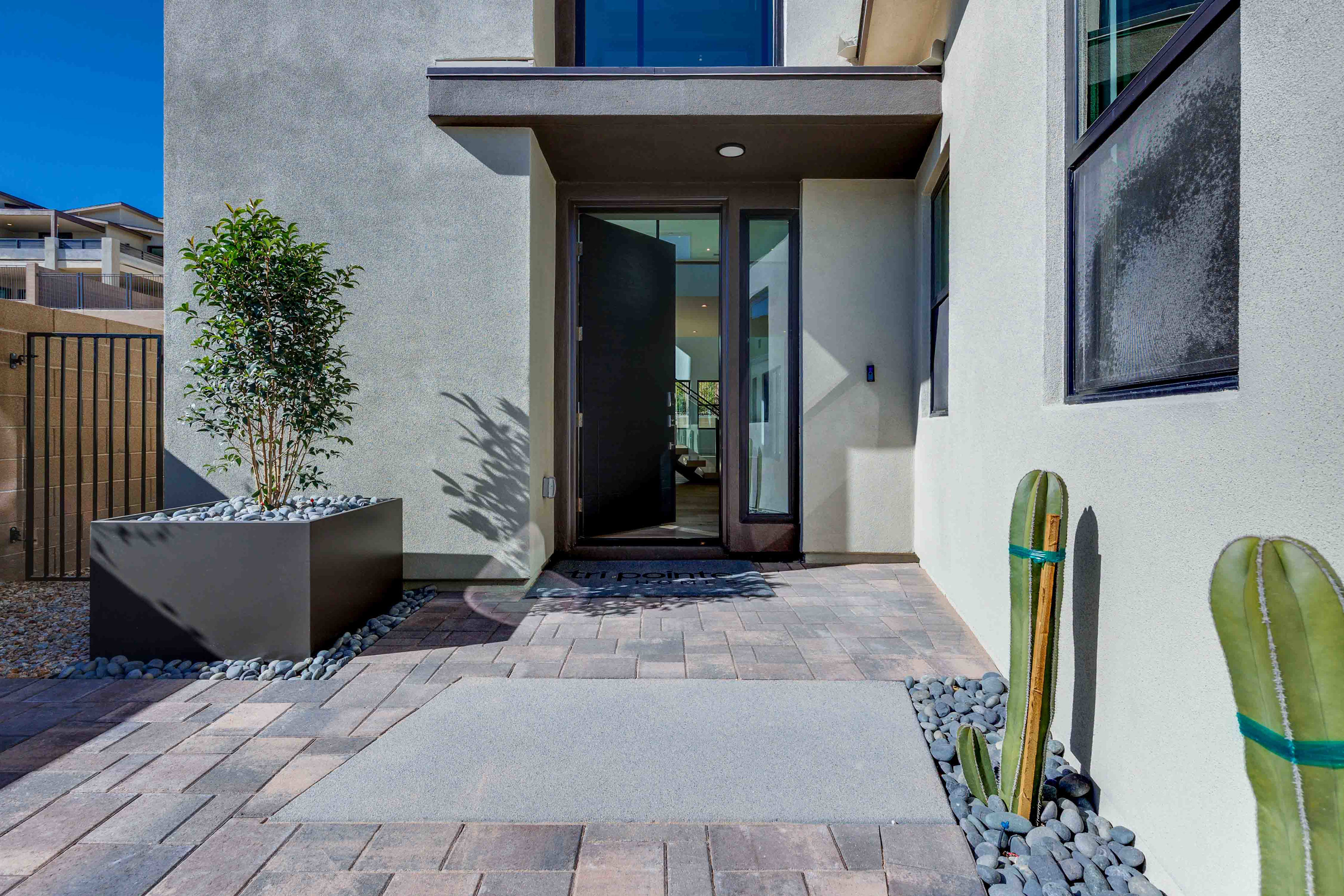 A modern, minimalist entryway with a paved walkway, potted plants, and a cactus sculpture leading to a glass-paneled front door against a backdrop of a white exterior wall.
