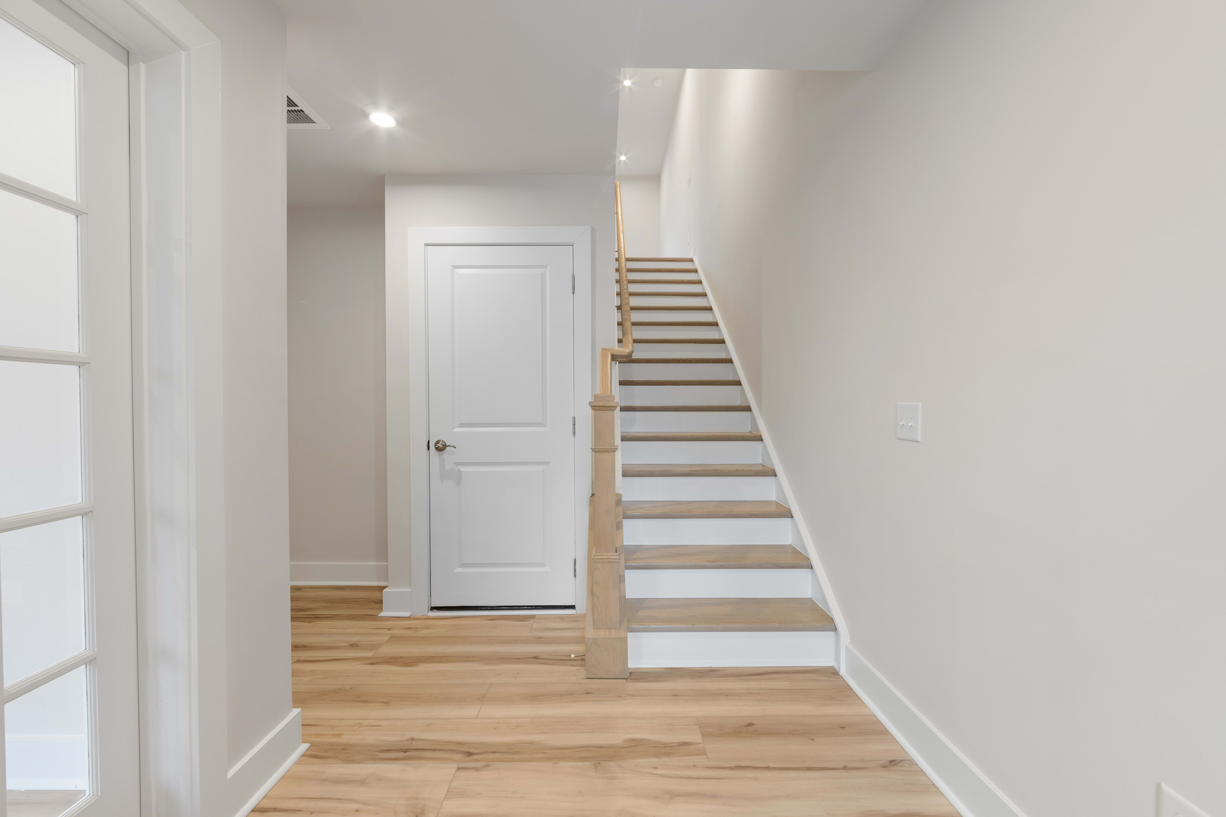 A bright and airy hallway with a white door, wooden stairs, and a hardwood floor leading to a well-lit space.