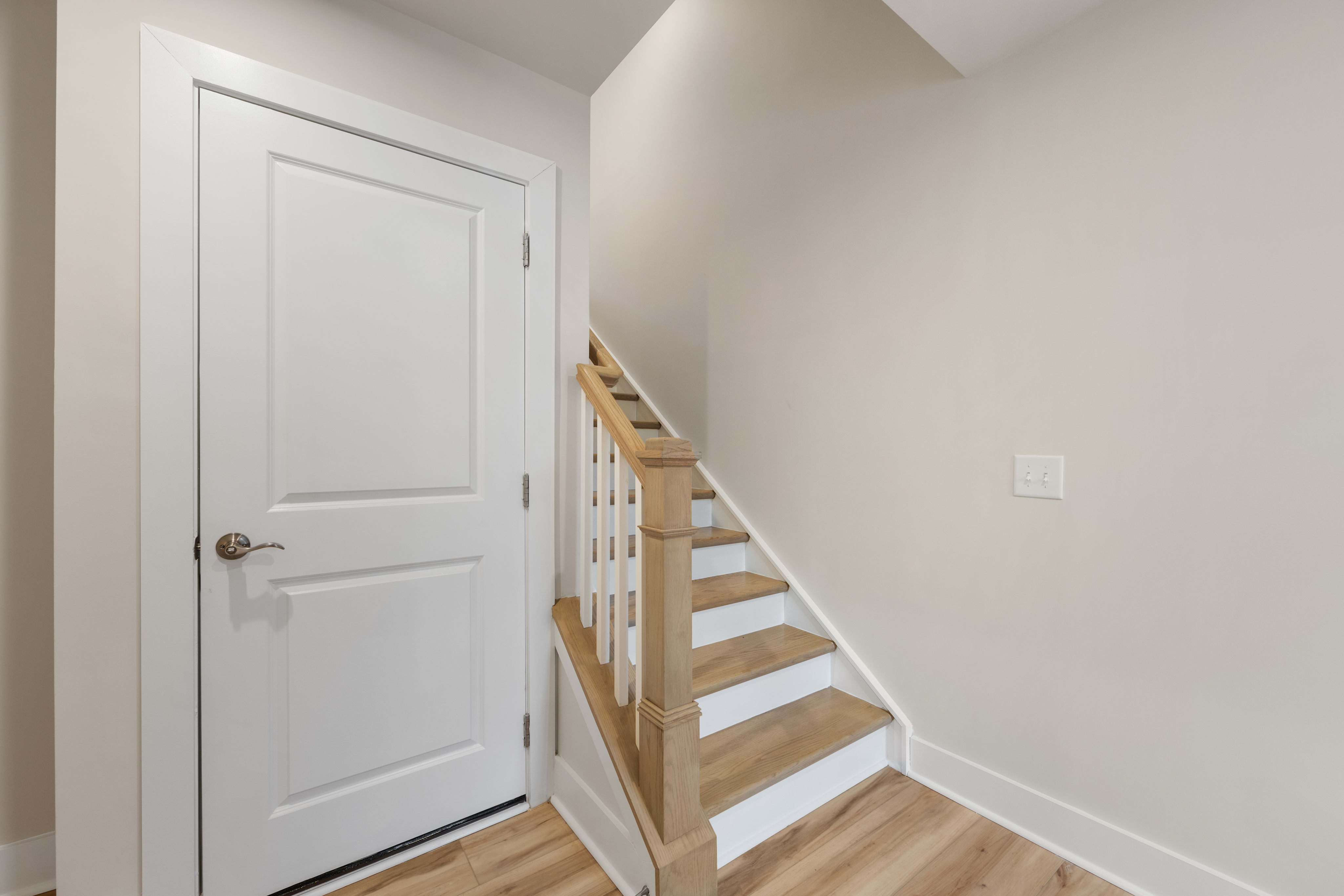 A white wooden door leads into a hallway with light-colored wooden stairs and a plain white wall in the background.