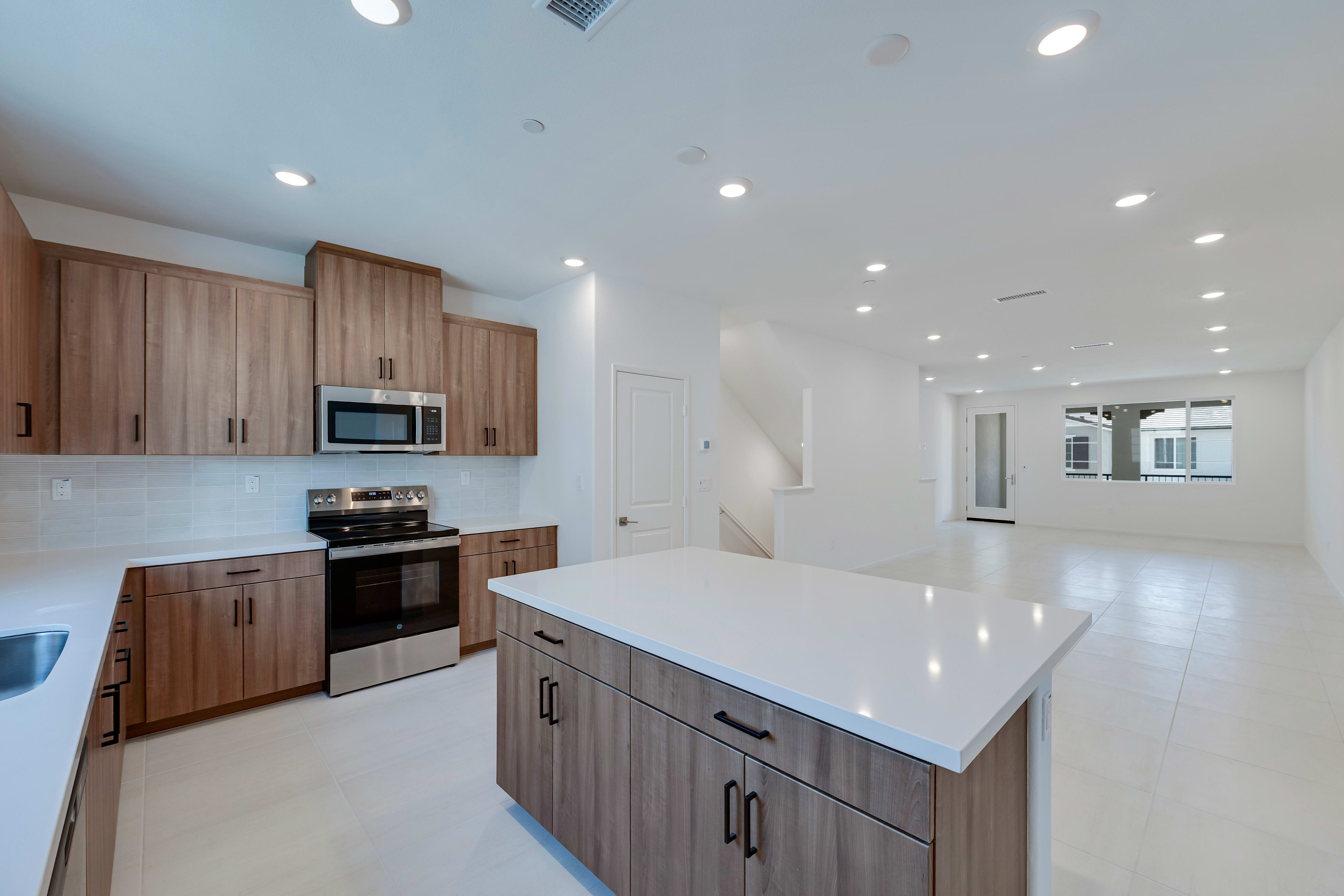 A modern, open-concept kitchen with wooden cabinets, white countertops, and stainless steel appliances, set against a bright and airy background.