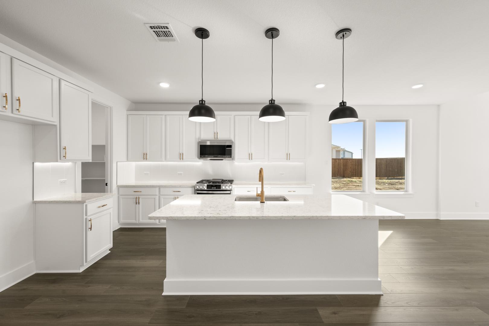 A modern, bright kitchen with white cabinets, a large island, and pendant lights hanging above it. The kitchen opens up to a view of the outdoors through a large window.